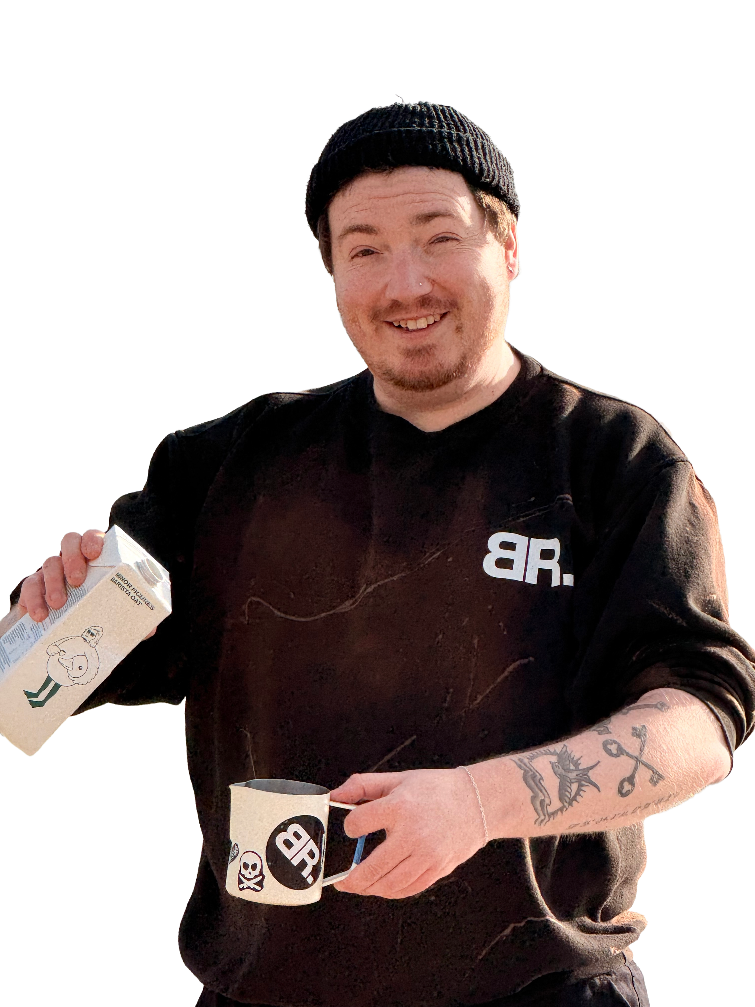 A person smiling, wearing a black beanie and black sweatshirt, holding a coffee cup and a carton of milk, with visible tattoos on their arms.