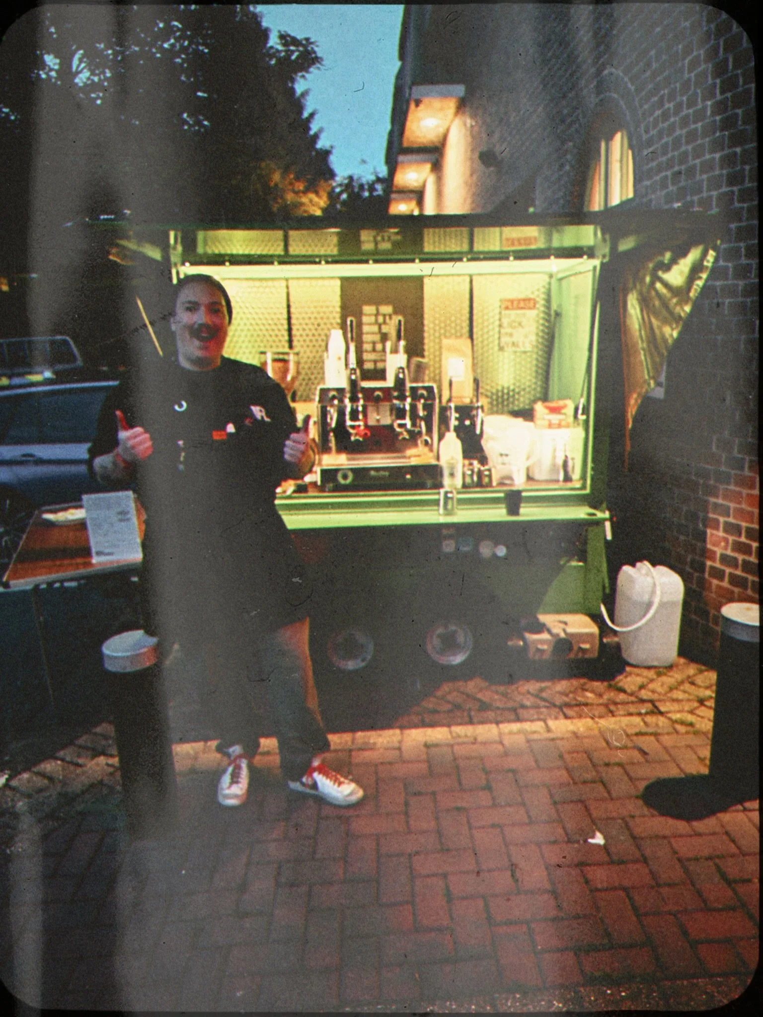 A man in a black shirt with a mustache, giving a thumbs-up, standing next to a green food truck serving coffee and snacks, with a brick sidewalk and trees in the background.