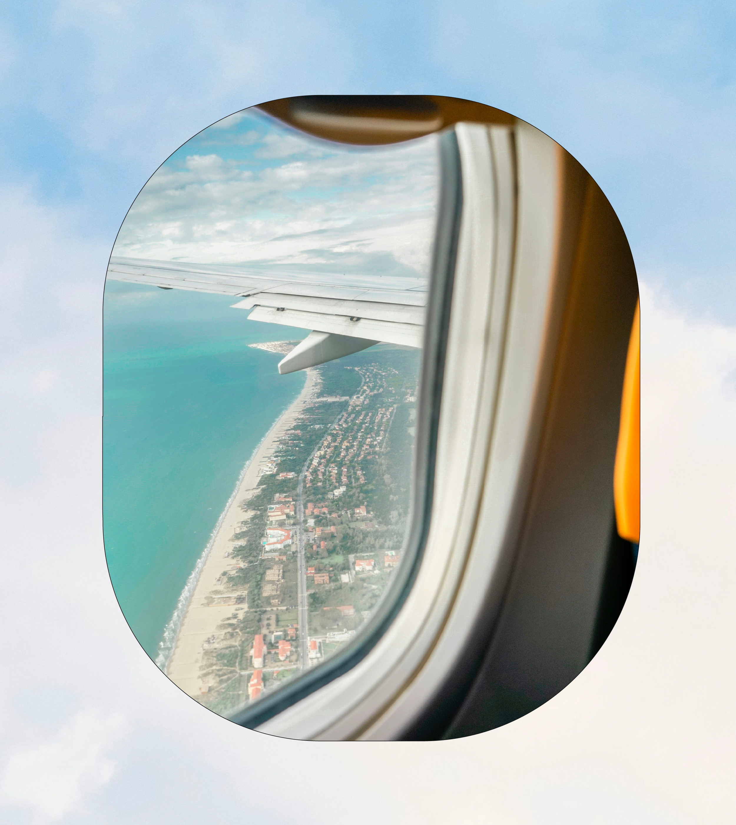 View from an airplane window showing a coastline with beach, residential area, and ocean, with a partly cloudy sky.