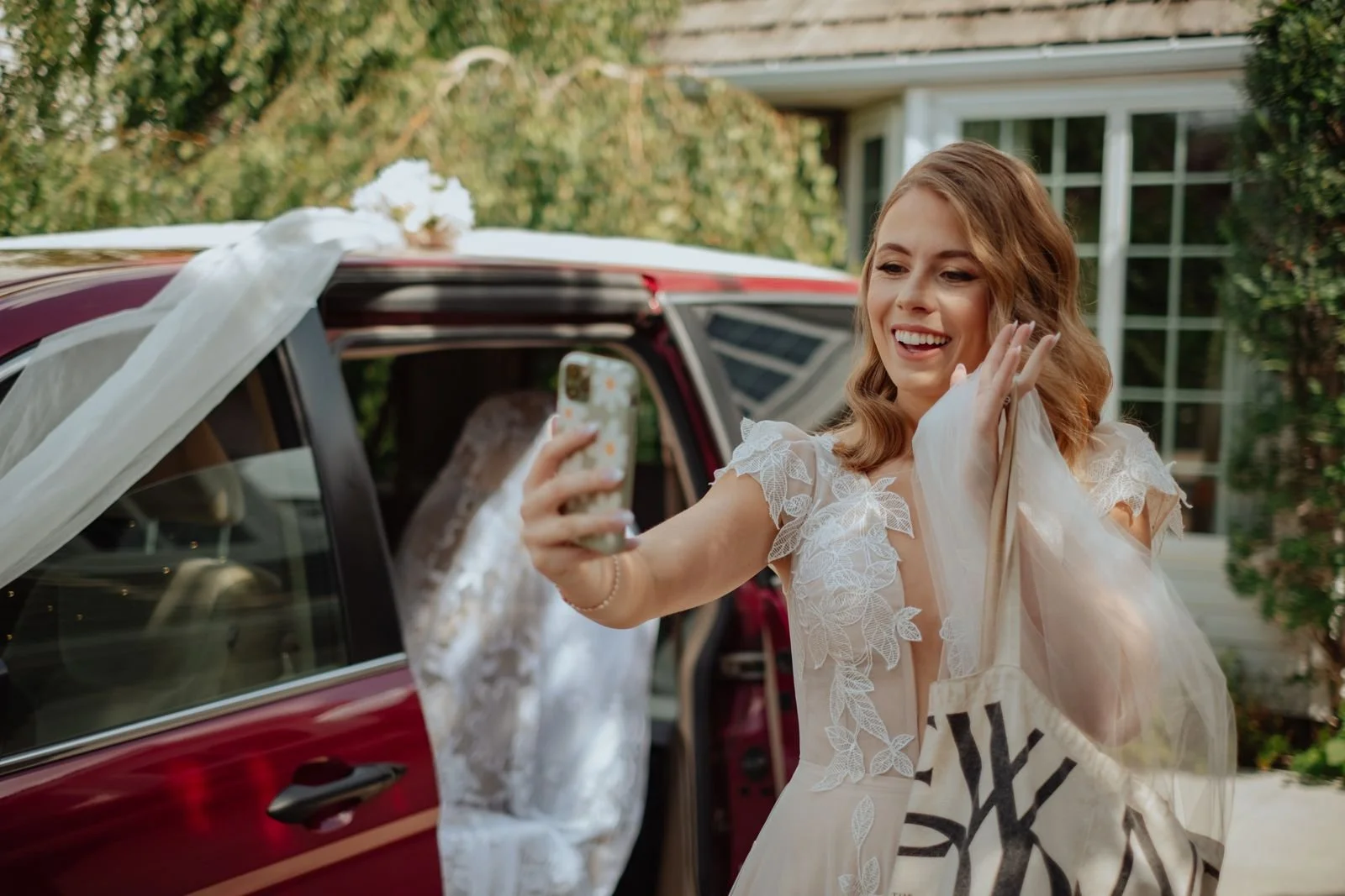 A woman in a white lace wedding dress taking a selfie outside near a red vehicle decorated for a wedding.