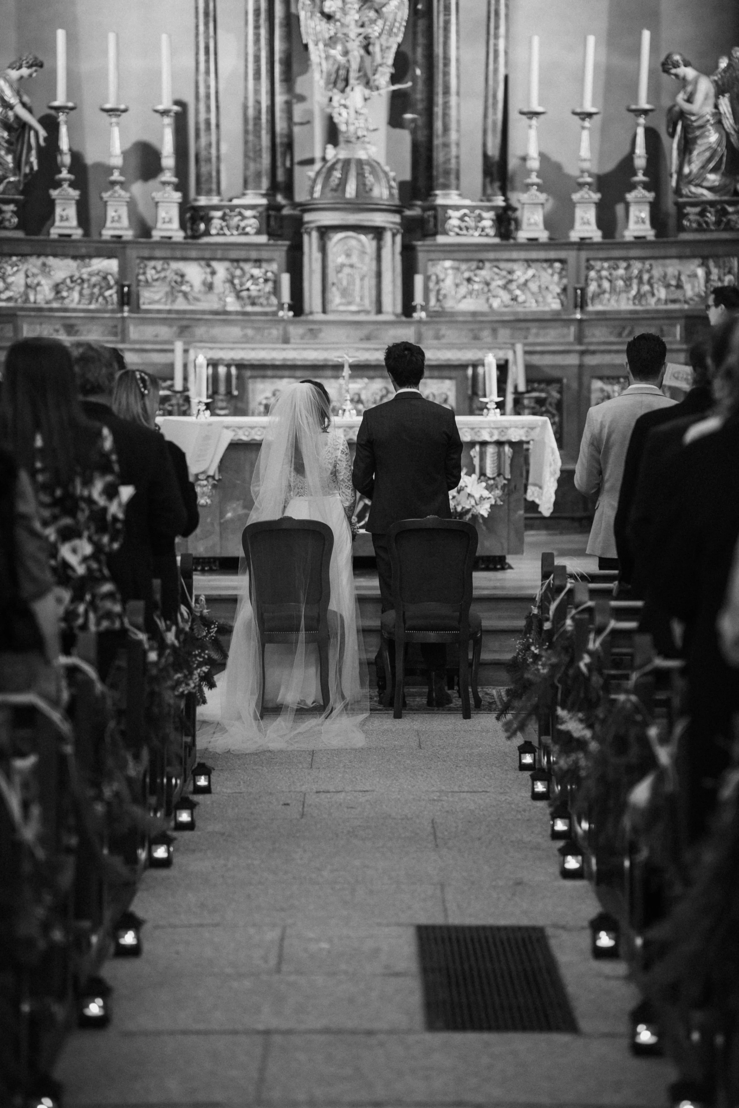 Black and white photo of a wedding ceremony inside a church with several guests seated, a bride and groom kneeling at the altar, and religious art and candles in the background.