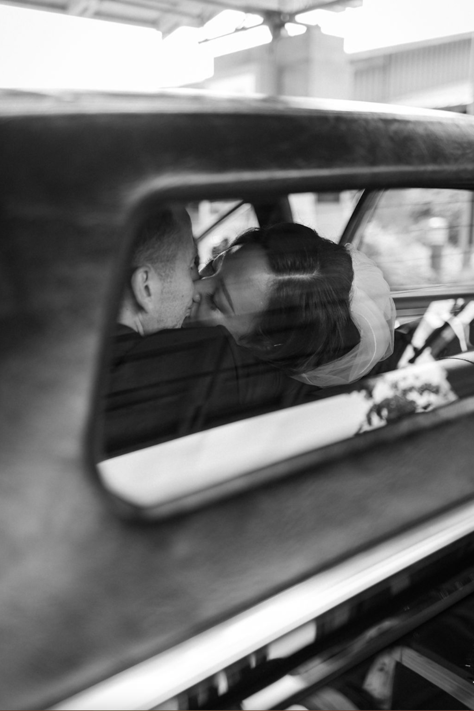A black-and-white photo of a couple sharing a kiss inside an old car, seen from through the back window.