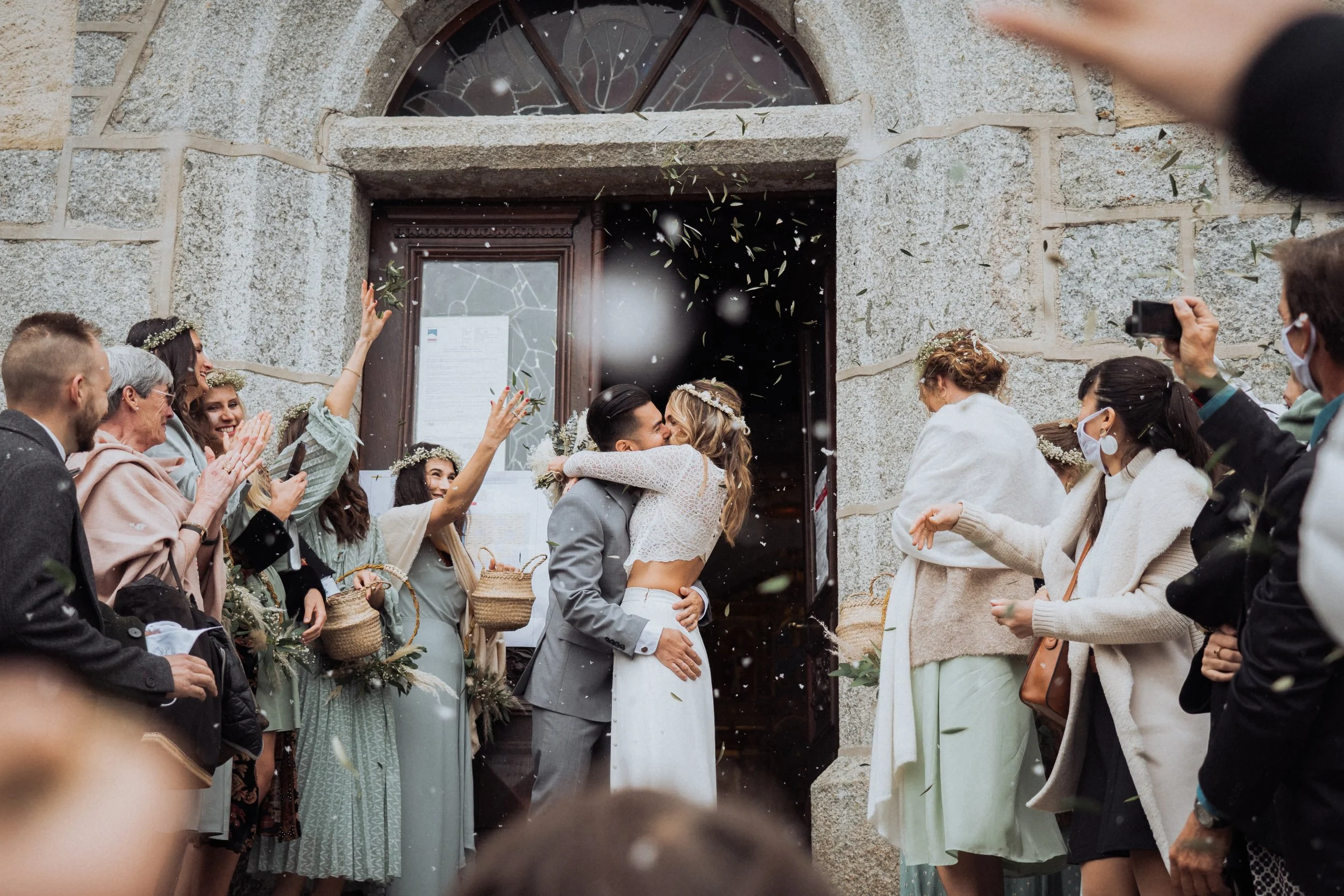 A newlywed couple kisses in front of a stone church door, surrounded by friends and family celebrating their wedding with confetti and joyful expressions.