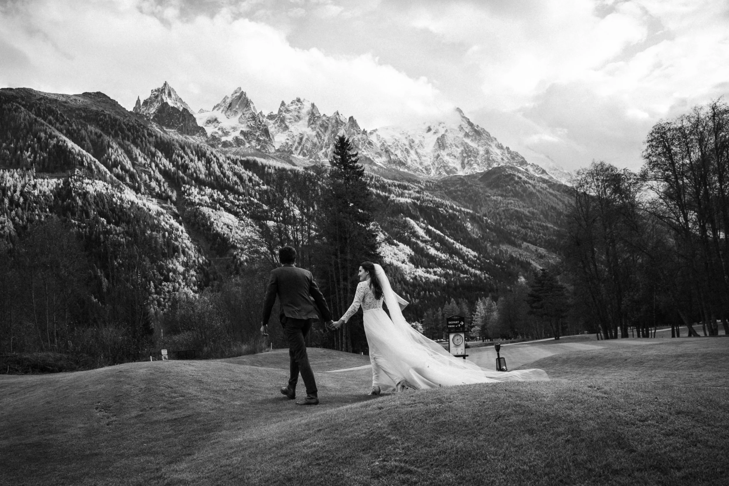 A bride and groom holding hands, walking on a grassy area with a mountain range in the background.