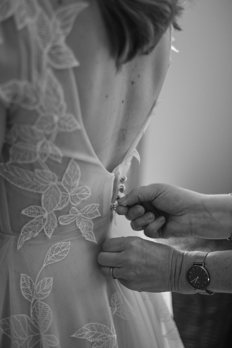 A person adjusting the buttons on the back of a wedding dress with lace leaf patterns, while another person's hands assist, wearing a watch.