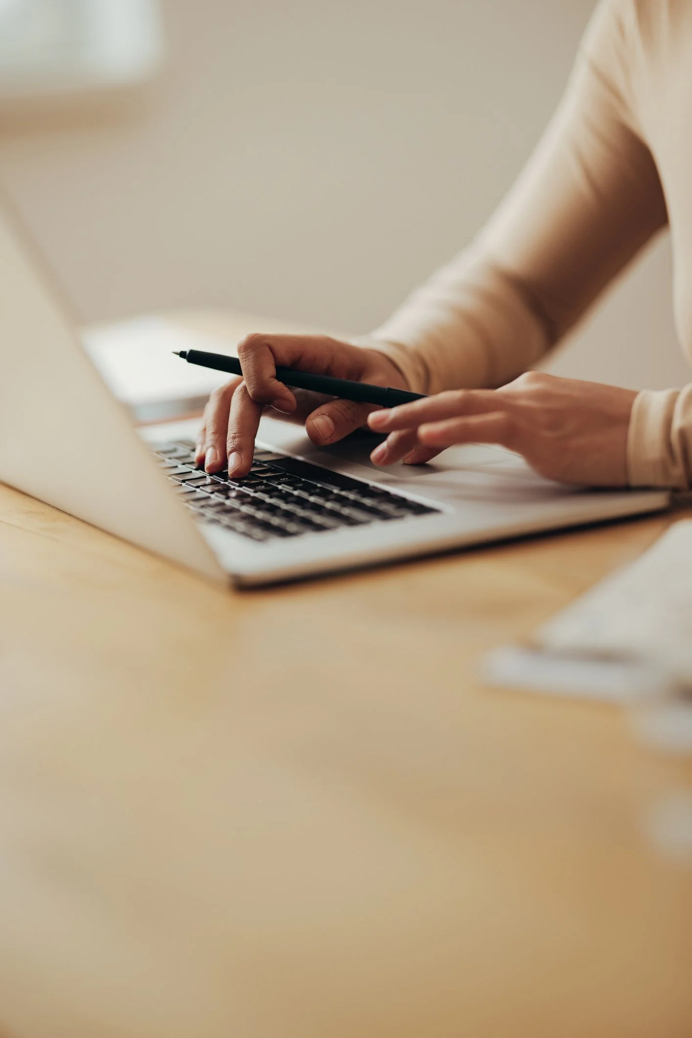 Close-up of a person's hands working on a laptop, holding a pen in one hand and typing on the keyboard with the other.