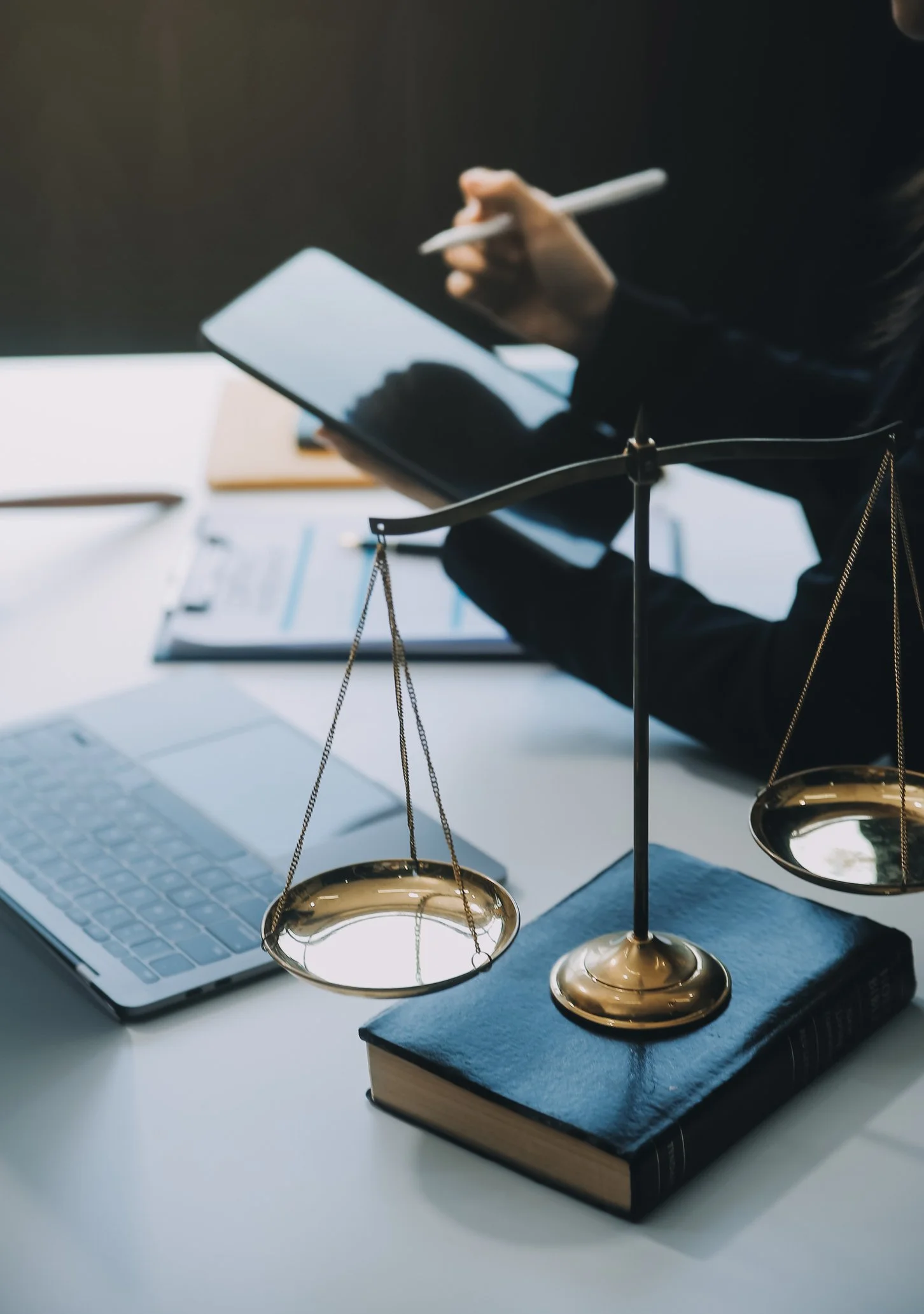 A person writing on a tablet at a desk with a laptop, a legal scale on a book, and documents.