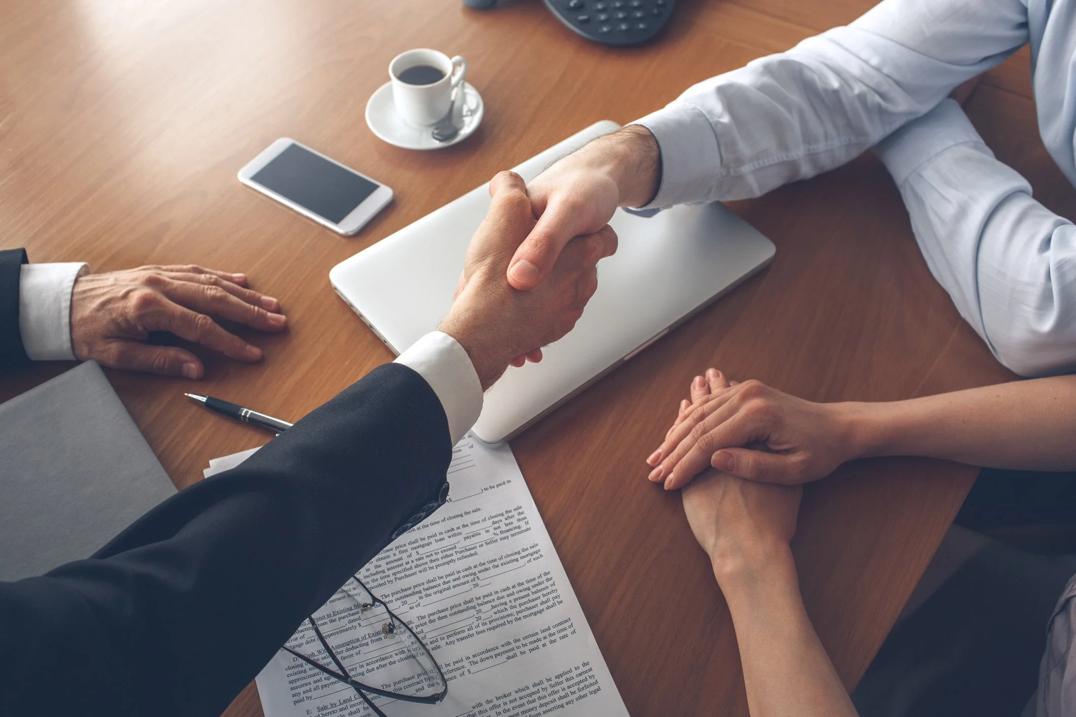 Two individuals shaking hands across a wooden table, with documents, a laptop, a smartphone, a cup of coffee, and a pen on the table.