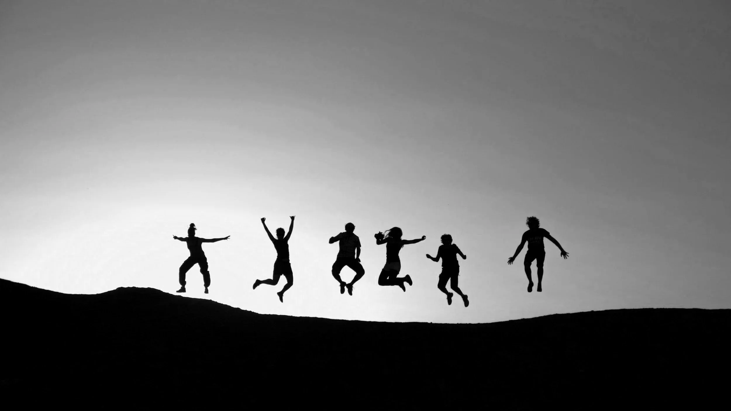 Group of children jumping and playing on a hill against a gray sky, silhouetted in black and white.