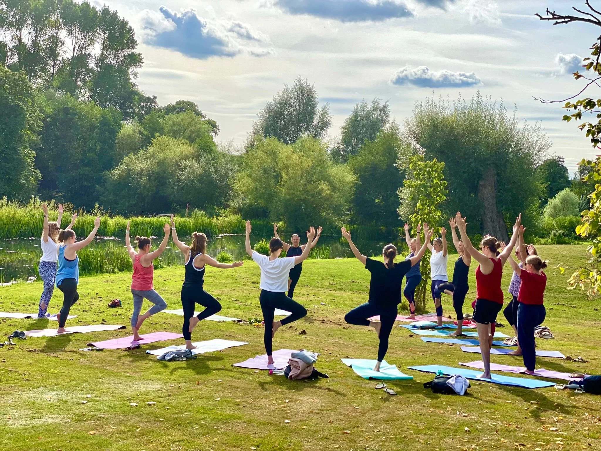 A group of people practicing yoga outdoors on a grassy area near a river, with trees and clouds in the sky.