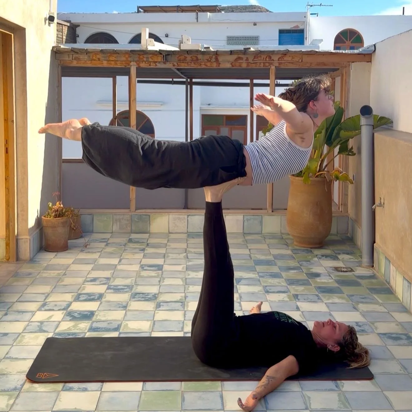 Two women practicing acro yoga on a rooftop terrace, with one lying on her back on a black yoga mat and supporting the other in a horizontal position with her legs and arm strength. The woman on the ground is wearing a black t-shirt and black pants, while the other woman is wearing a striped tank top and dark pants. The terrace has a tiled floor and potted plants, with a background of white buildings and blue sky.
