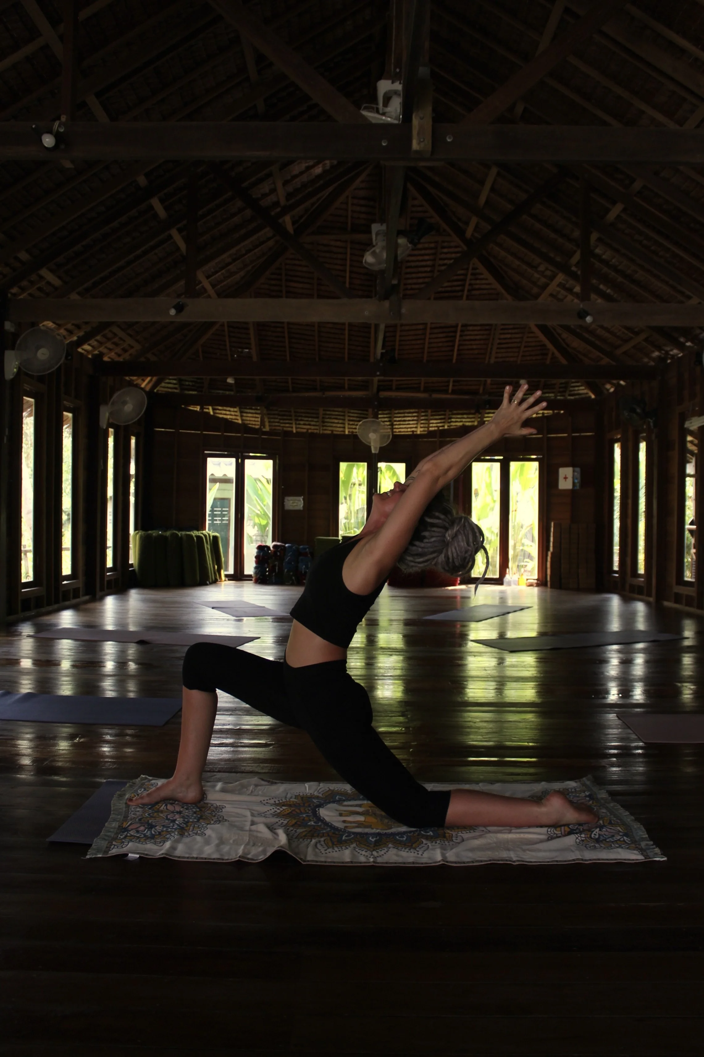 A woman practicing yoga in a wooden, open-air studio with large windows, sunlight, and greenery outside, performing a low lunge pose with arms extended upward.