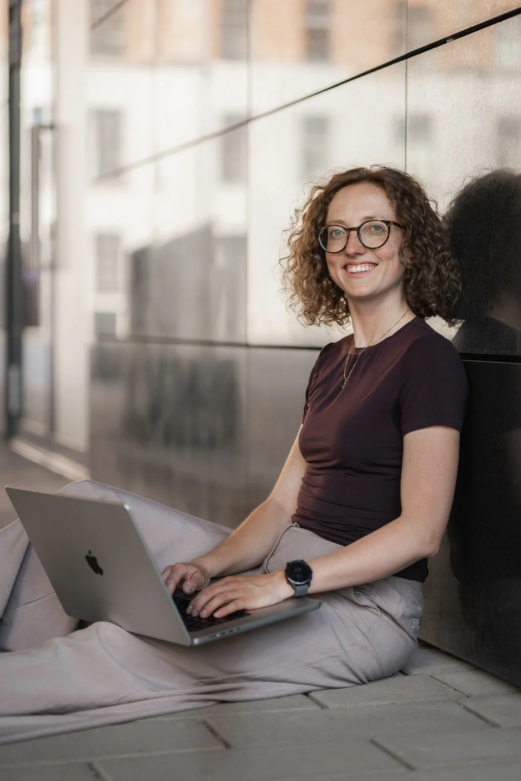 Junge Frau mit Brille sitzt auf dem Boden an einer Wand mit einem Laptop auf dem Schoß, lächelt in die Kamera.