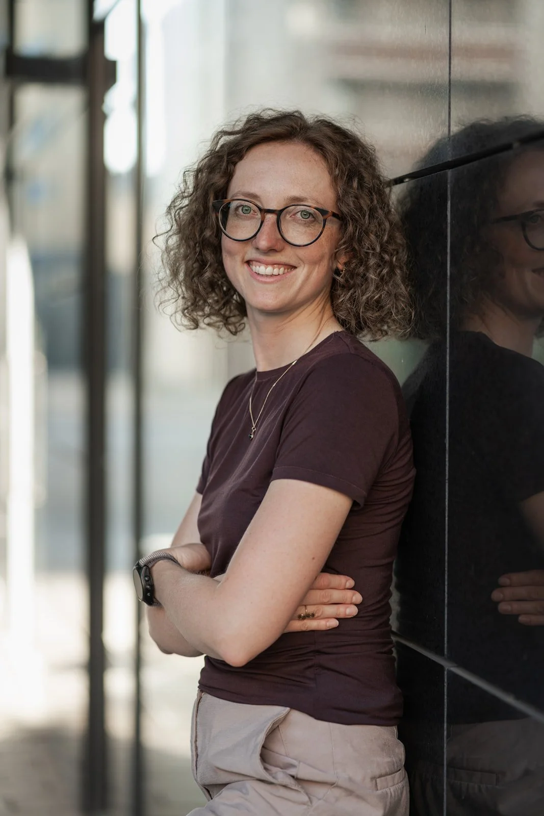Junge Frau mit Brille, braunem T-Shirt und beige Hose, lächelnd, gegen eine schwarze Wand gelehnt, im Hintergrund verschwommene urbane Umgebung.