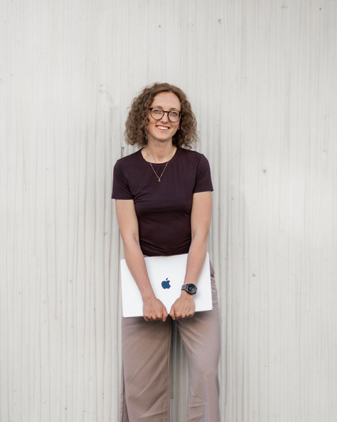 Frau mit Brille, lockigem Haar, dunklem T-Shirt, beige Hosen, hält einen MacBook mit Apple-Logo vor sich, steht vor einer hellen Wand.