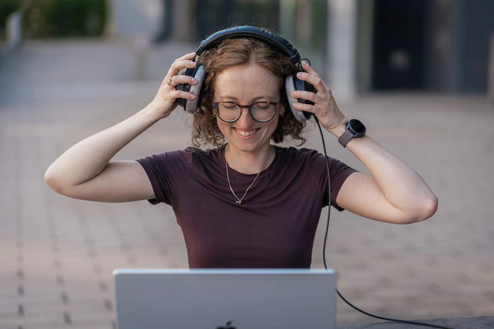 Eine lachende junge Frau trägt Kopfhörer, steht draußen vor einem Laptop, und scheint Musik oder einen Podcast zu genießen.
