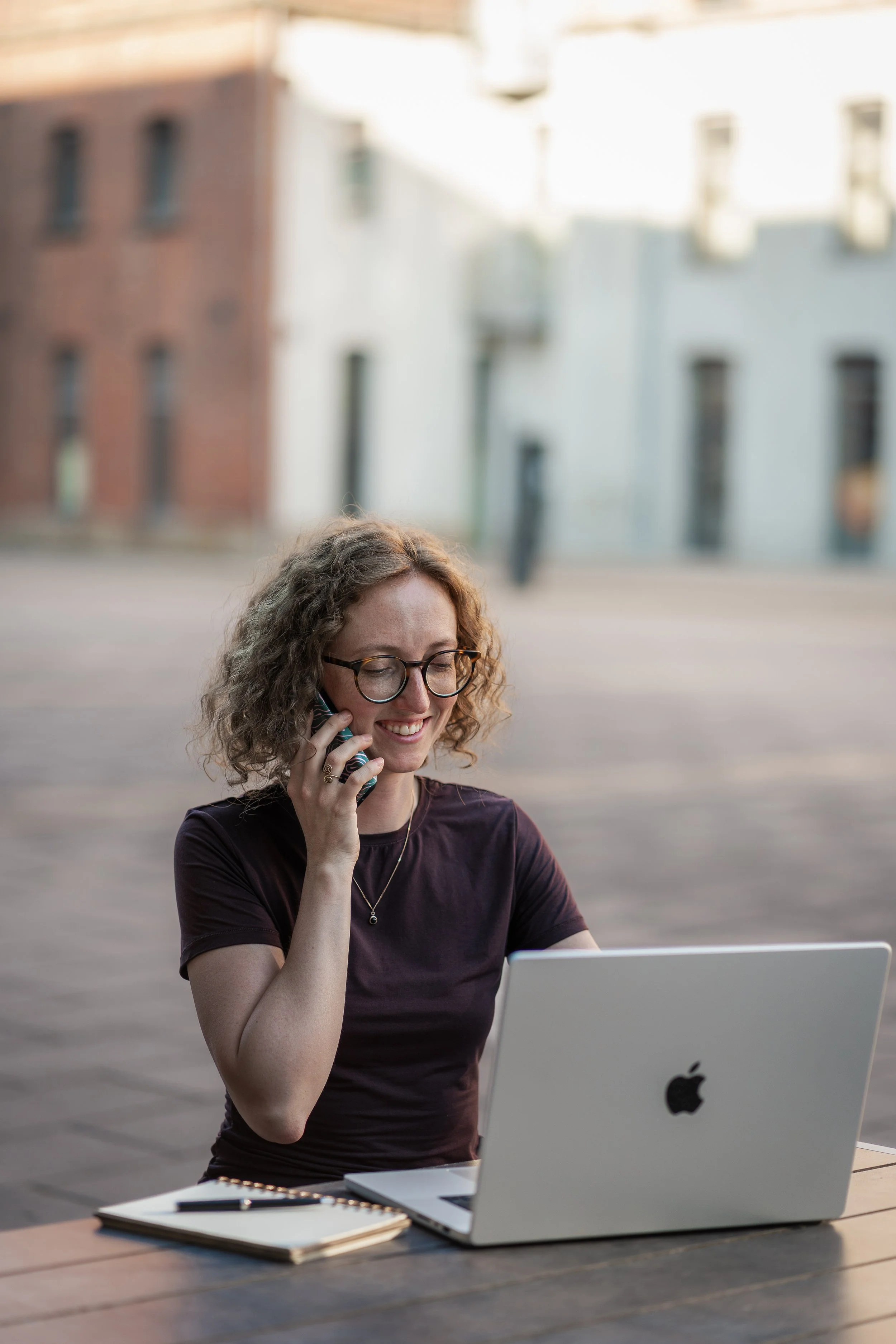 Junge Frau mit Brille, teilt Lächeln, spricht am Handy, sitzt an einem Tisch mit einem Laptop und Notizbuch im Freien.