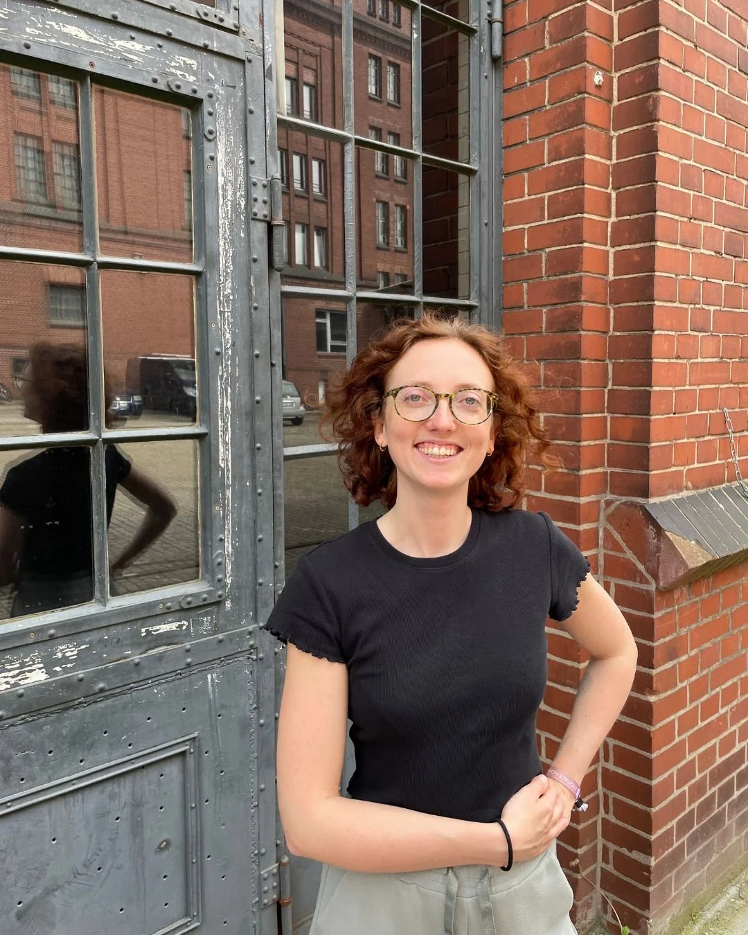 Virtuelle Assistenz Mira Kullmann vor einem alten Fenster und roten Backsteinmauern in einer urbanen Umgebung.
