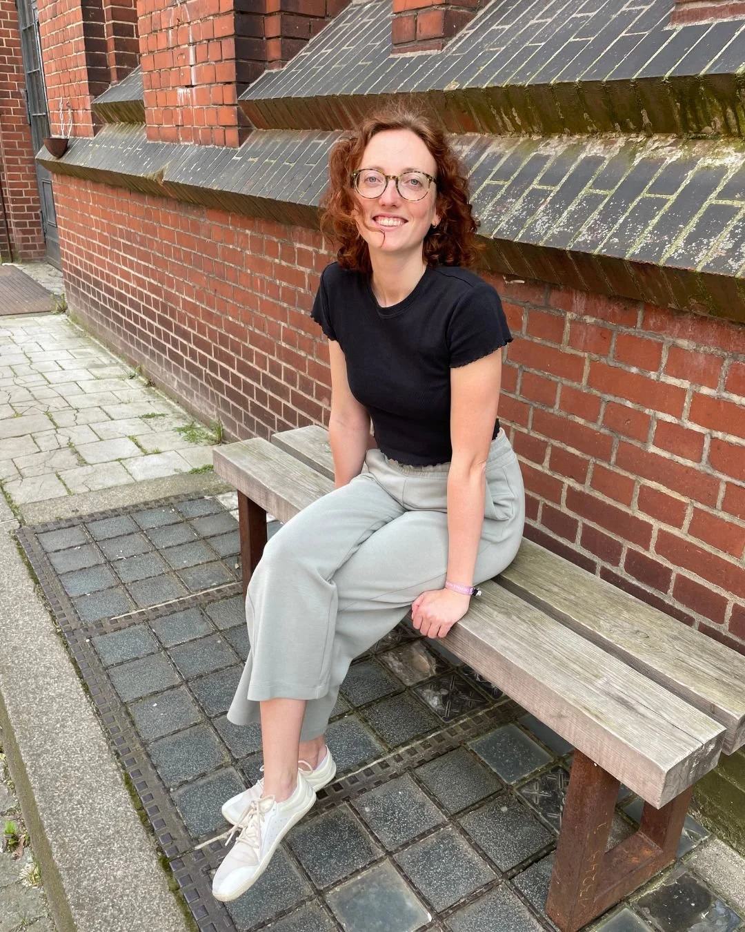 Virtuelle Assistenz Mira Kullmann sitzt auf einer Holzbank vor einer roten Backsteinmauer.