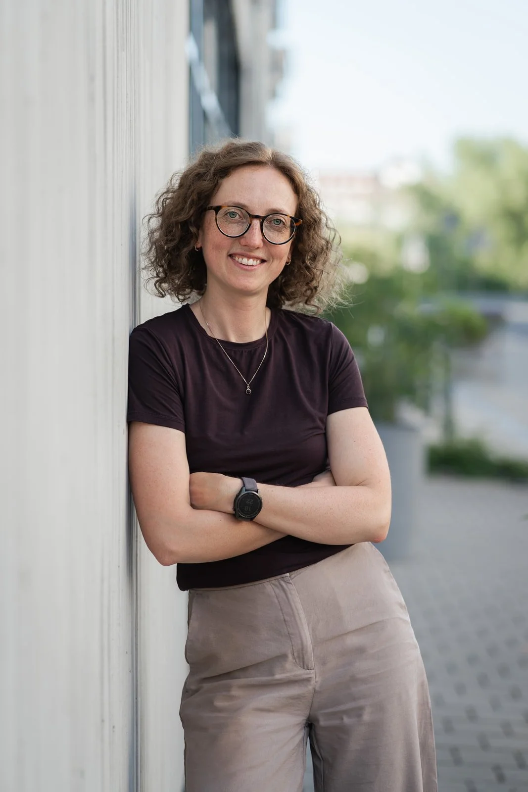 Eine lächelnde Frau mit Brille, kurzen lockigen Haaren, schwarzen T-Shirt, beige Hosen, Armbanduhr, Kette, steht draußen vor einer Wand, mit einem freundlichen Ausdruck, im Hintergrund Bäume und Himmel.