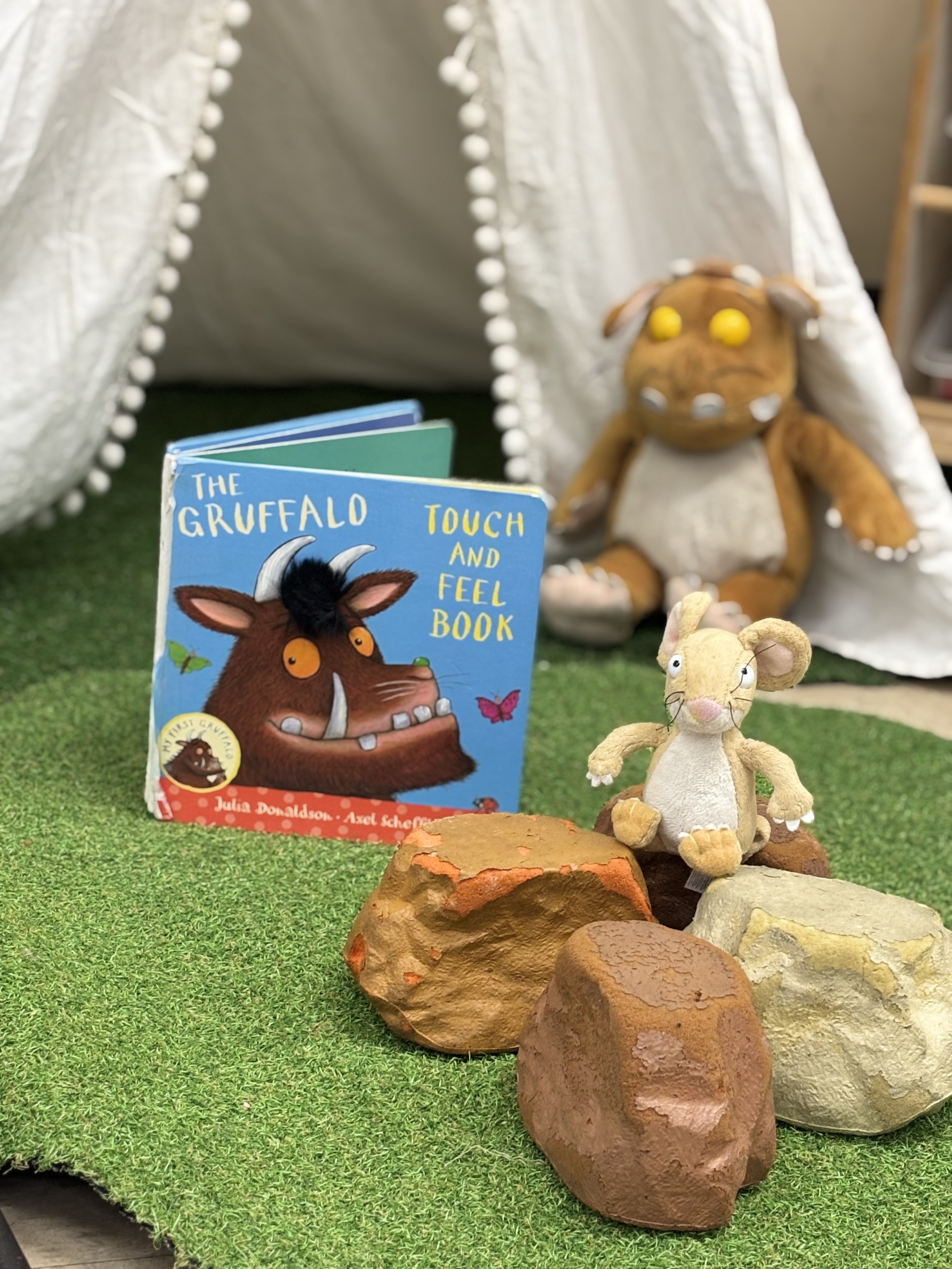 A small plush mouse toy, a children's book titled 'The Gruffalo Touch and Feel Book,' and several painted rocks are arranged on a green turf-like surface with a plush Gruffalo toy and a plush mouse toy in the background at Pop-Up Day Nursery
