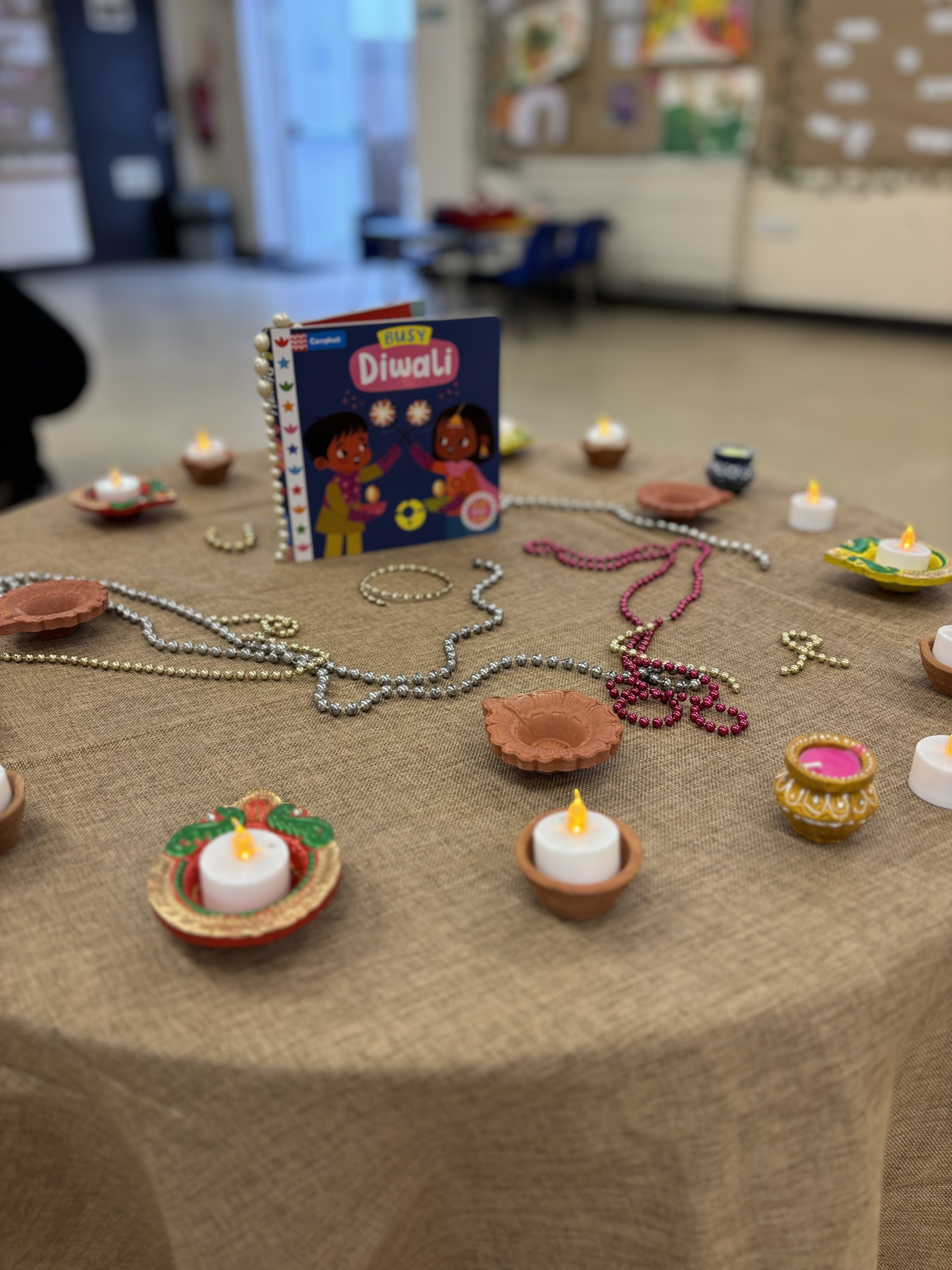 Diwali celebration setup with small oil lamps, colorful jewelry, and a children's book titled 'Busy Diwali' on a table, in a classroom or similar setting at Pop-Up Day Nursery