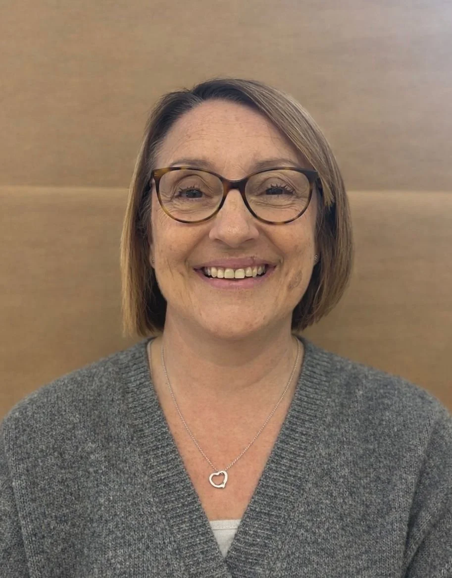 A smiling woman with glasses in front of a plain light-colored background, wearing a black hoodie.