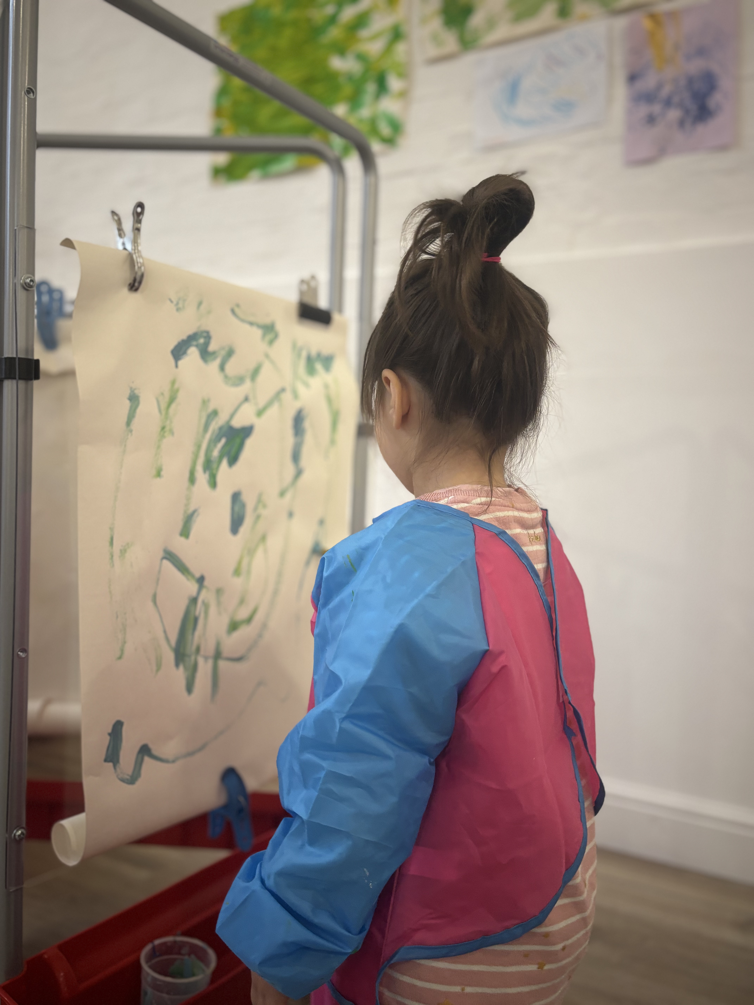 A young girl with long dark hair tied up in a half-up hairstyle, wearing a pink and striped top with a blue and pink protective apron, stands in front of an easel with a large sheet of paper displaying green and blue children's artwork.