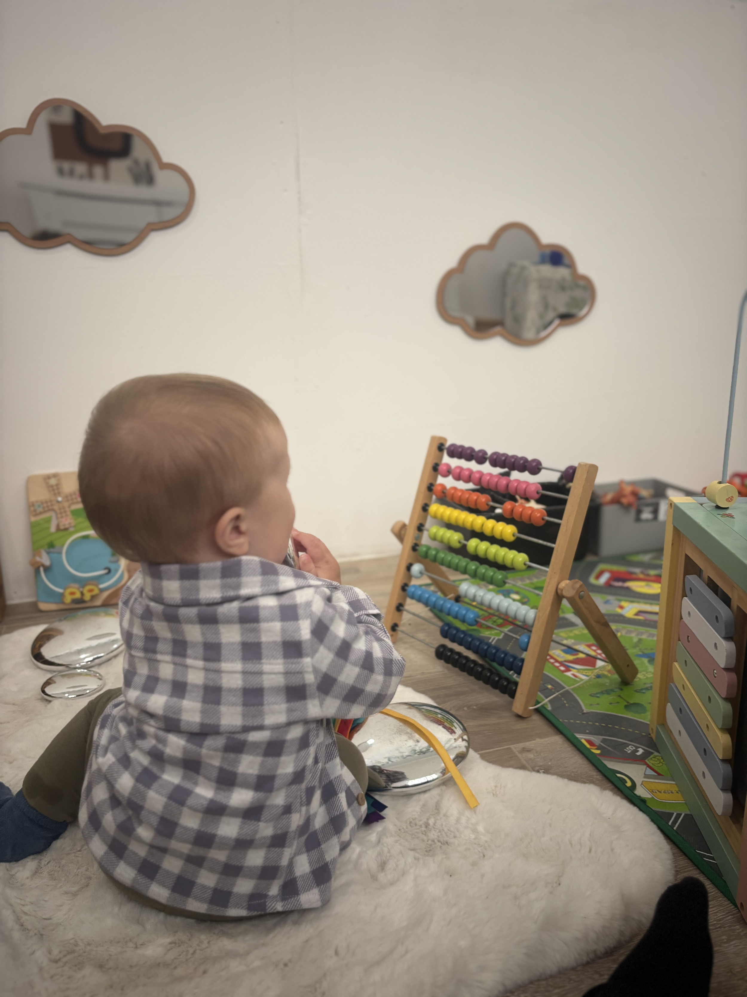 A young child sitting on a cream-colored rug, surrounded by toys, watching an abacus and other game items in a playroom with cloud-shaped mirrors on the wall at Pop-Up Day Nursery.