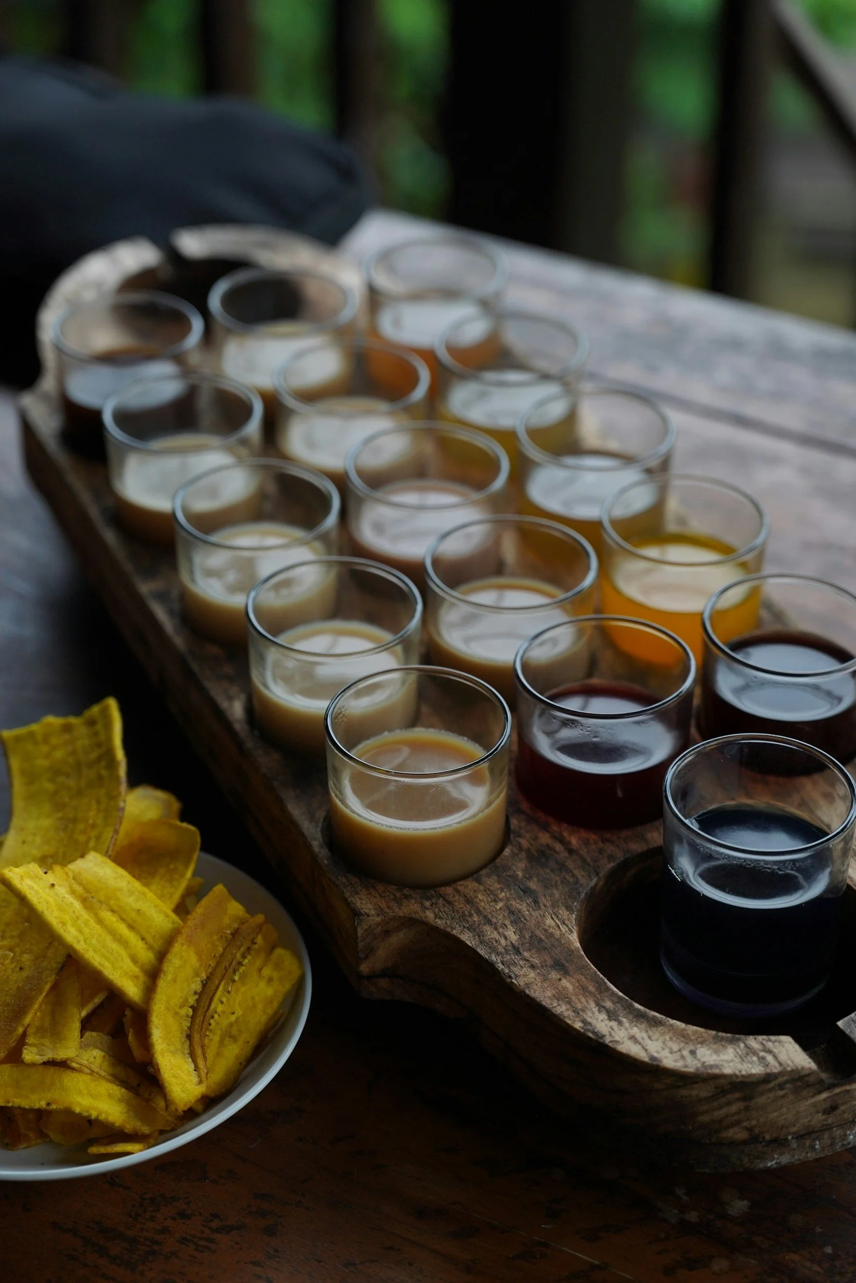A wooden flight paddle with multiple small glasses of different types of beer and a white plate of plantain chips on a wooden table.