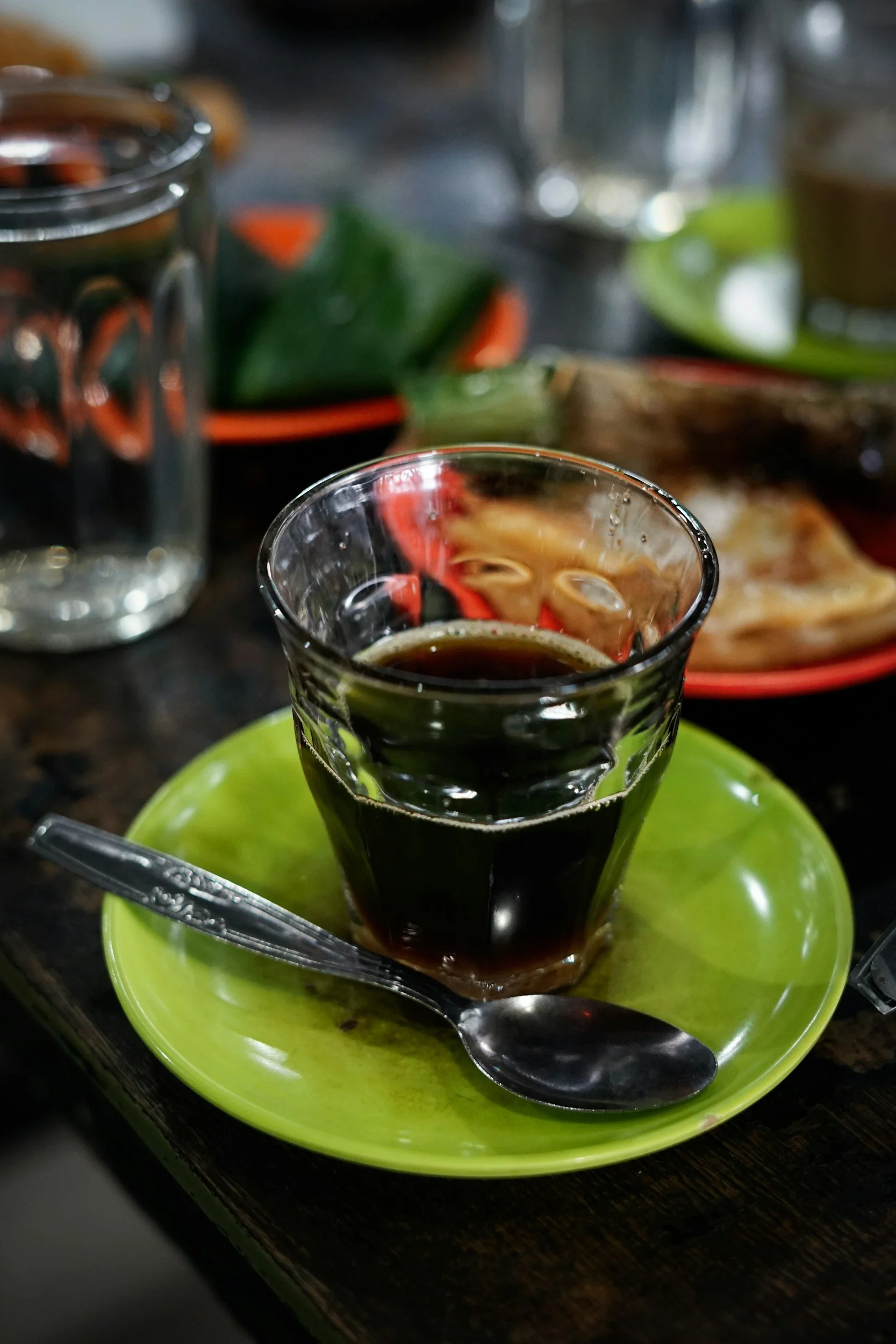 A glass of black coffee on a bright green saucer with a metal spoon, surrounded by dishes and jars, on a dark wooden table.
