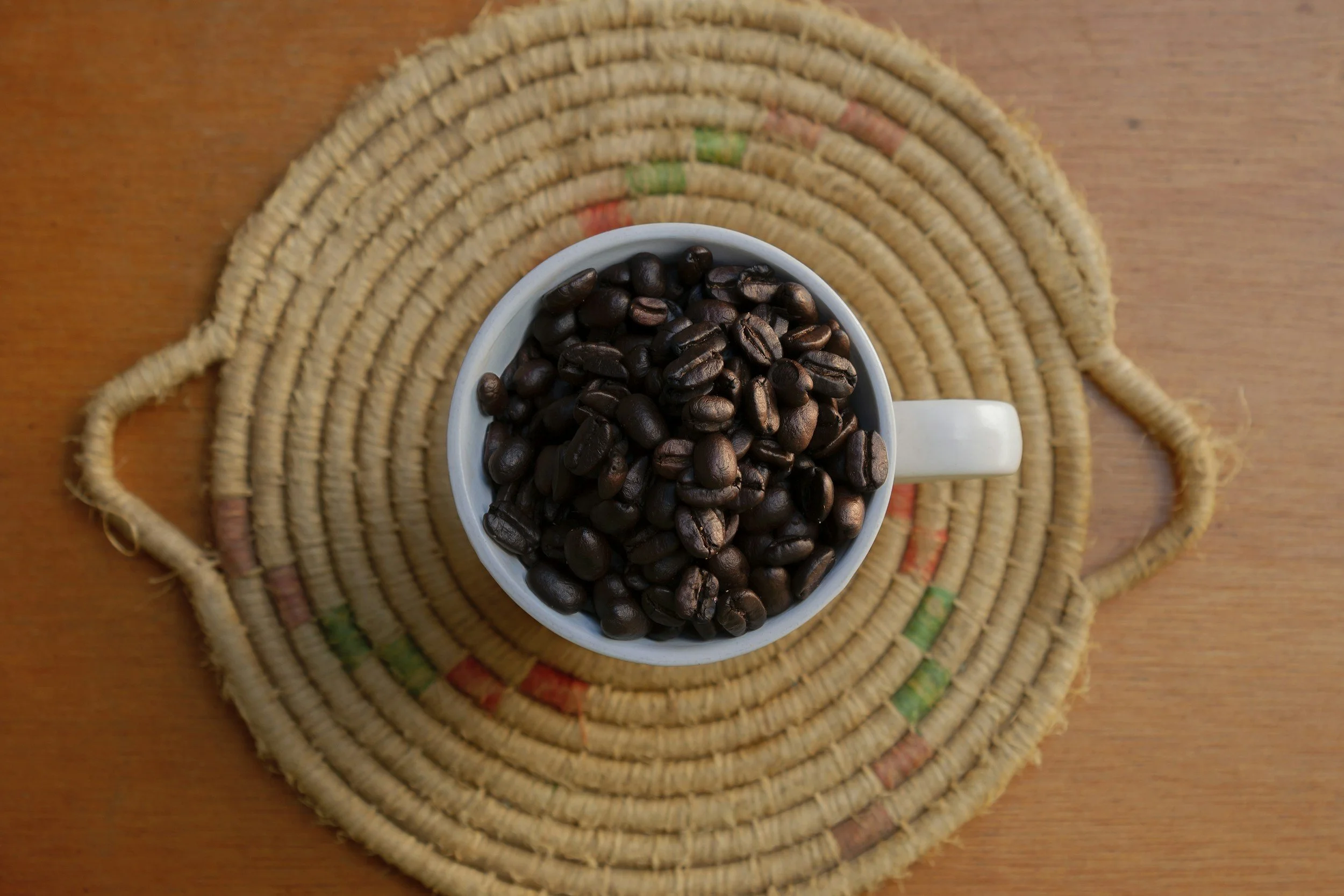 A white coffee mug filled with dark roasted coffee beans on a woven round placemat on a wooden table.