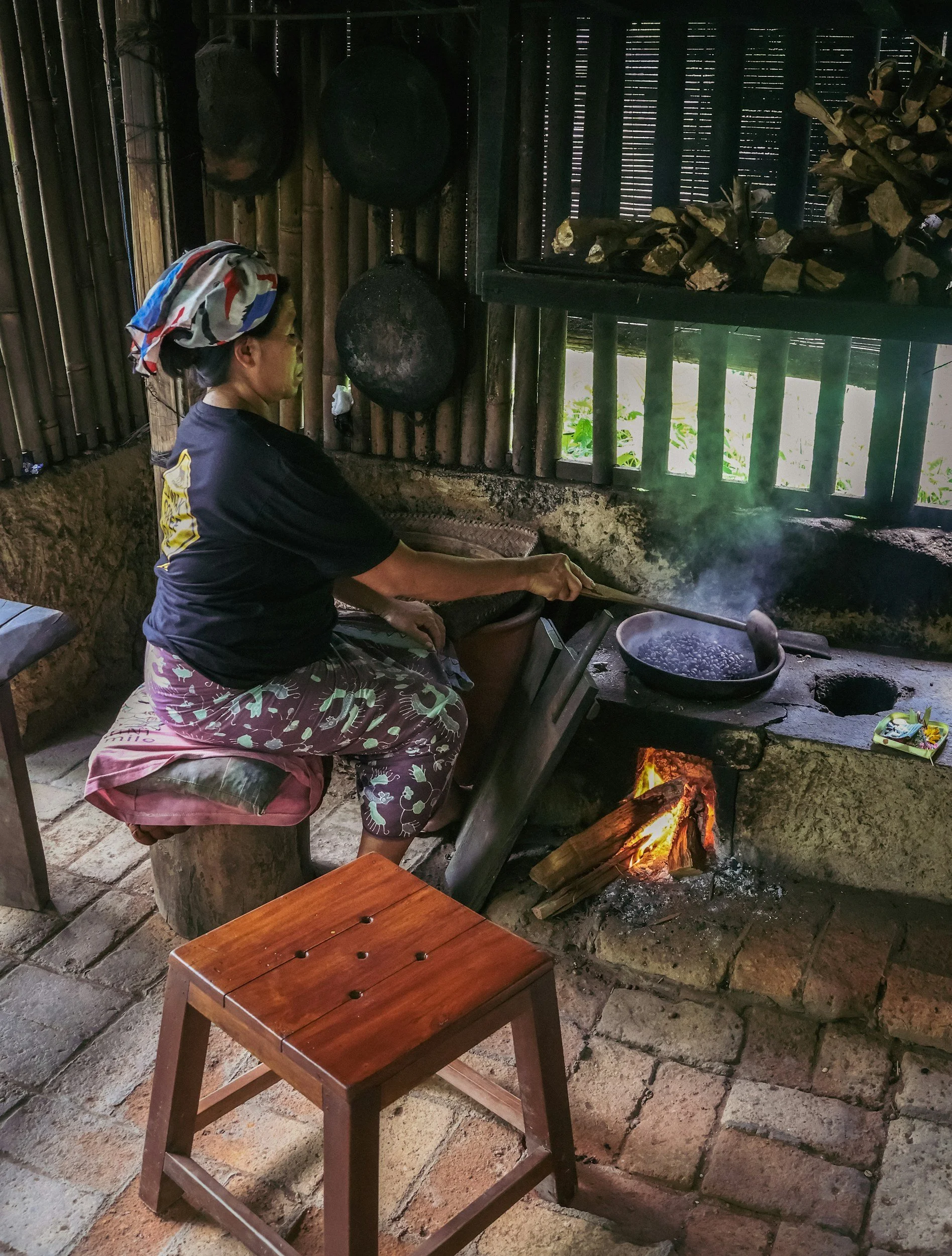 A woman cooking over an open fire in a traditional kitchen with a wood stove, using a frying pan with steam rising from it, and wearing a colorful headscarf and casual clothing.