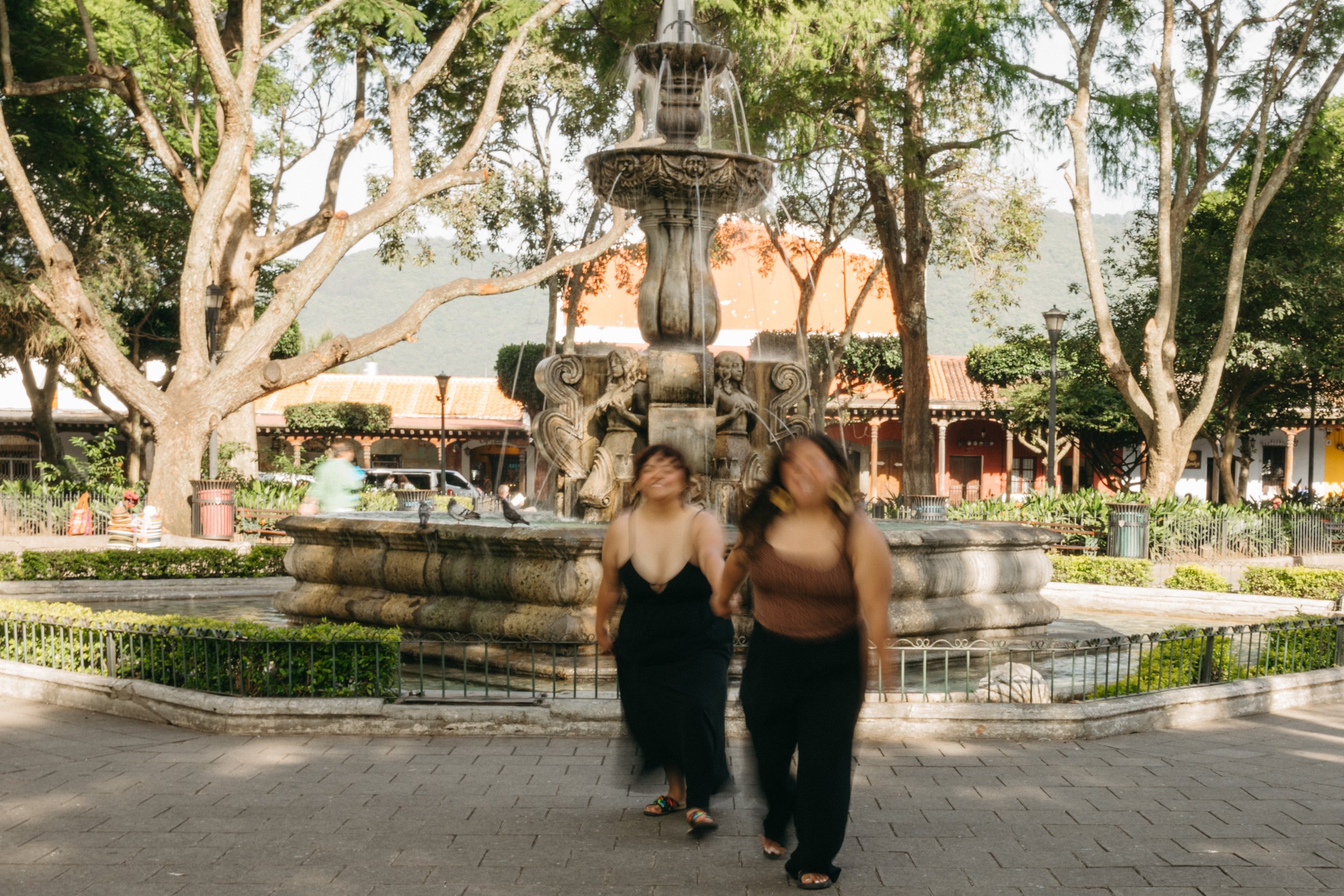 Lesbian couple walking hand in hand in front of a fountain in Antigua Guatemala Central Park (part of Diana Alvarado’s lifestyle and LGBTQ+ couple photography in Guatemala).