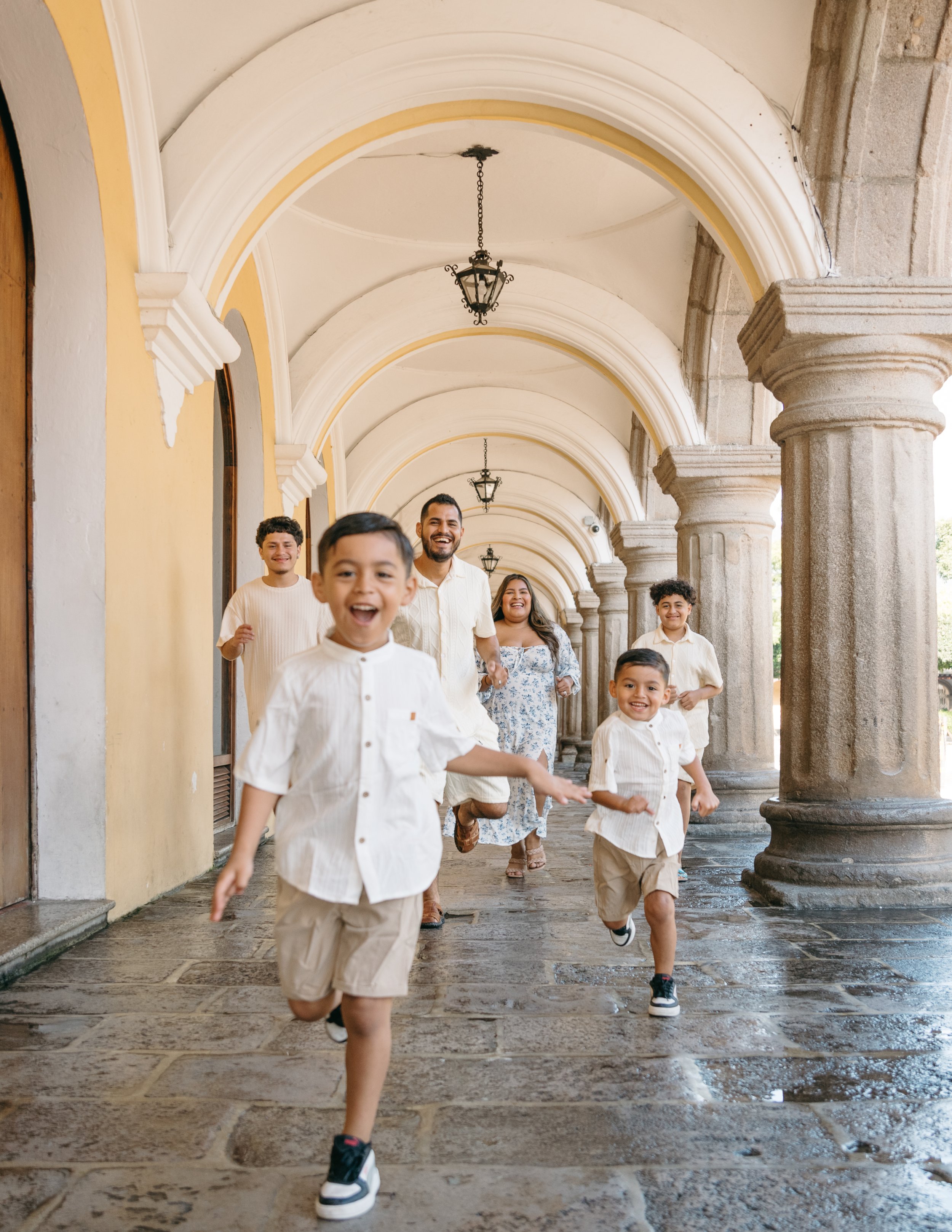 Family session with smiling children and two adults running at Palacio de los Capitanes building captured in a joyful lifestyle portrait in Antigua Guatemala.

