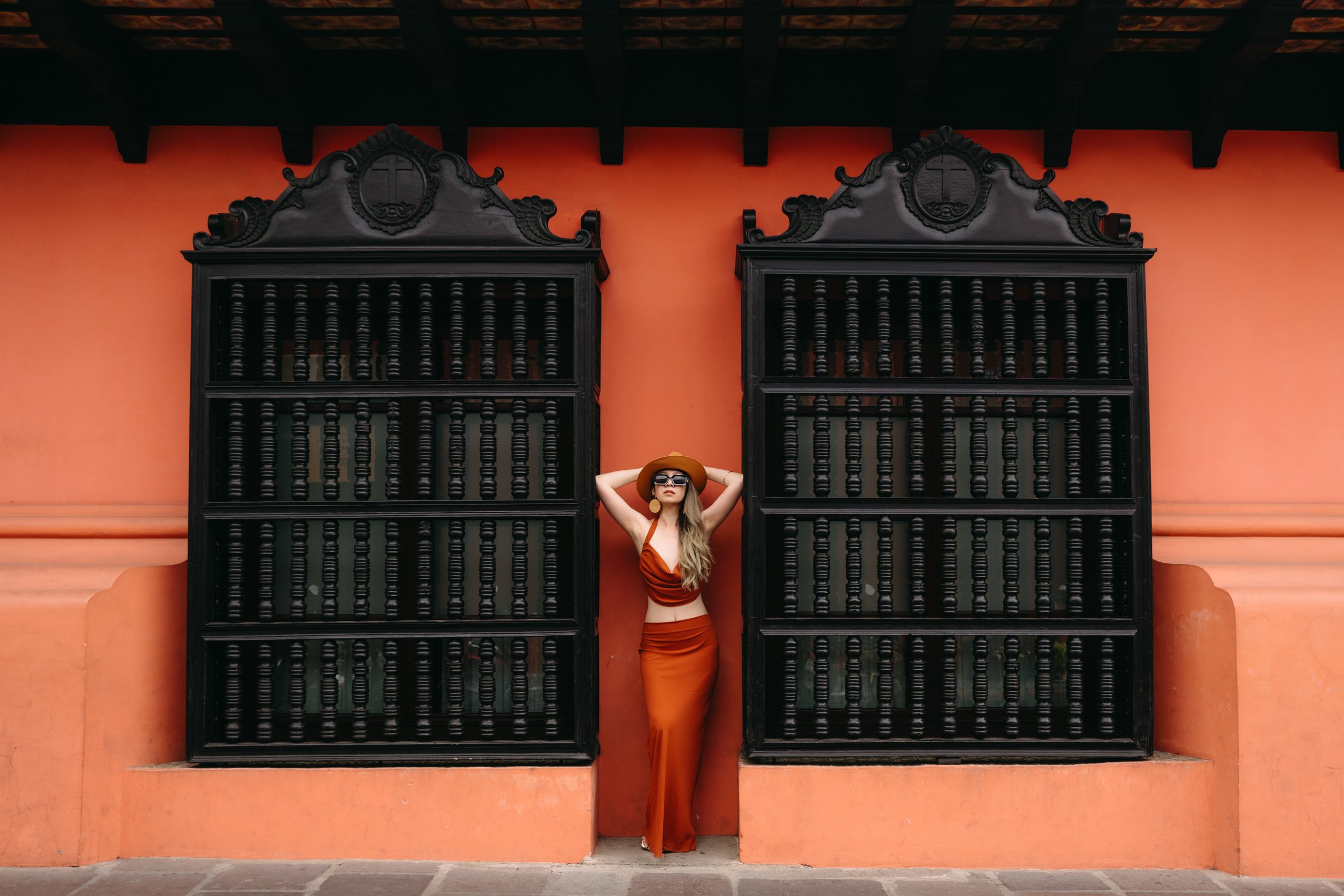 Woman in an orange dress and hat posing for a lifestyle portrait between two large black wooden windows in the colorful colonial city of Antigua, Guatemala.