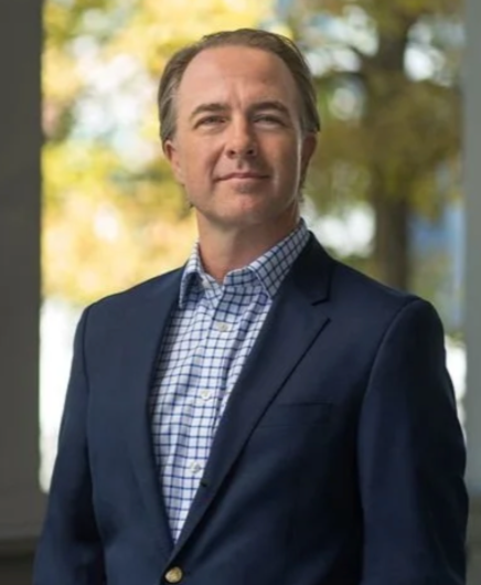A man in a suit and checkered shirt standing outdoors with trees in the background.