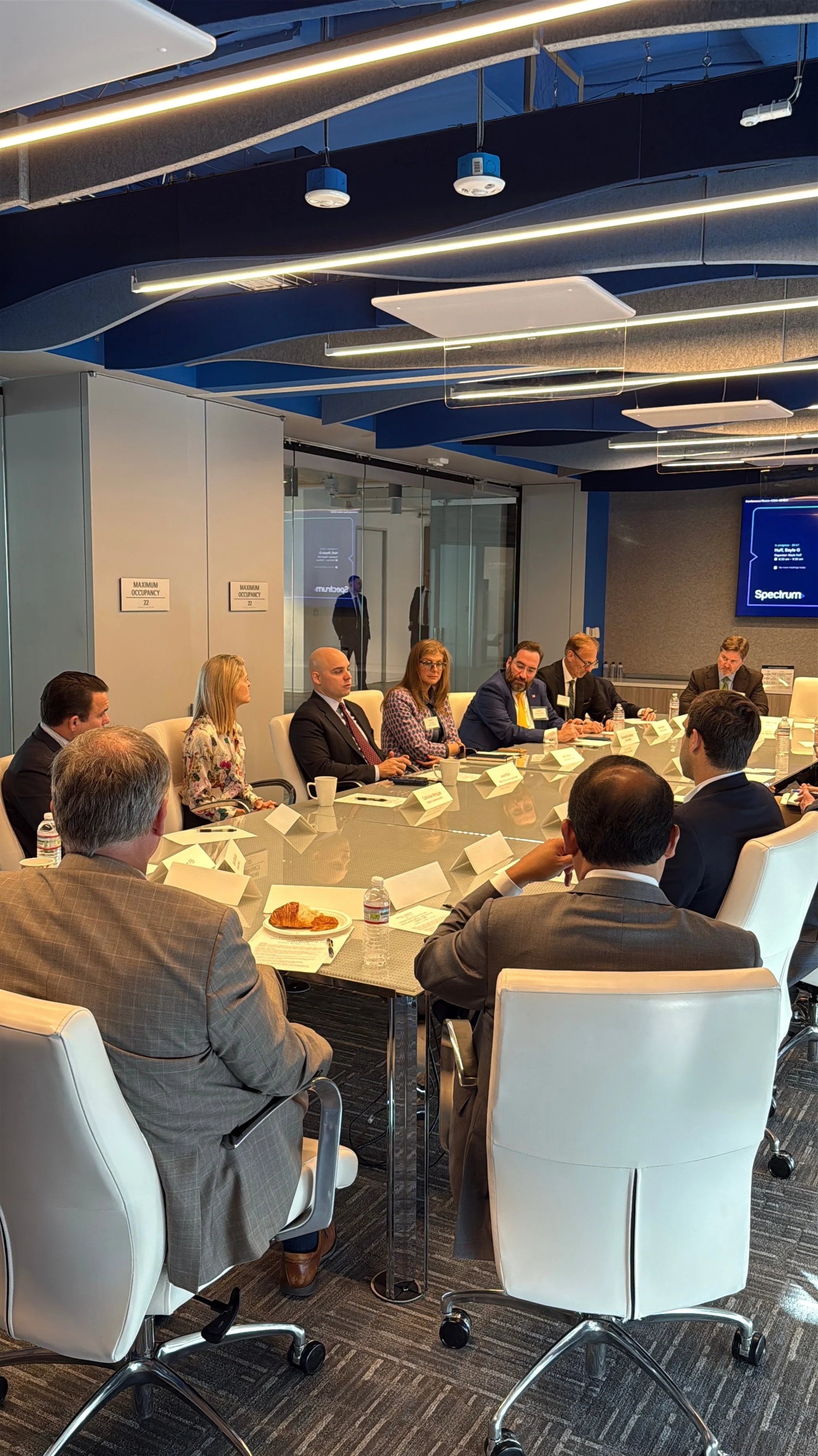 An AGP meeting taking place in a modern conference room with a glass wall. Several people in formal attire are seated around a large, oval conference table. Some have papers, laptops, and water bottles in front of them. 