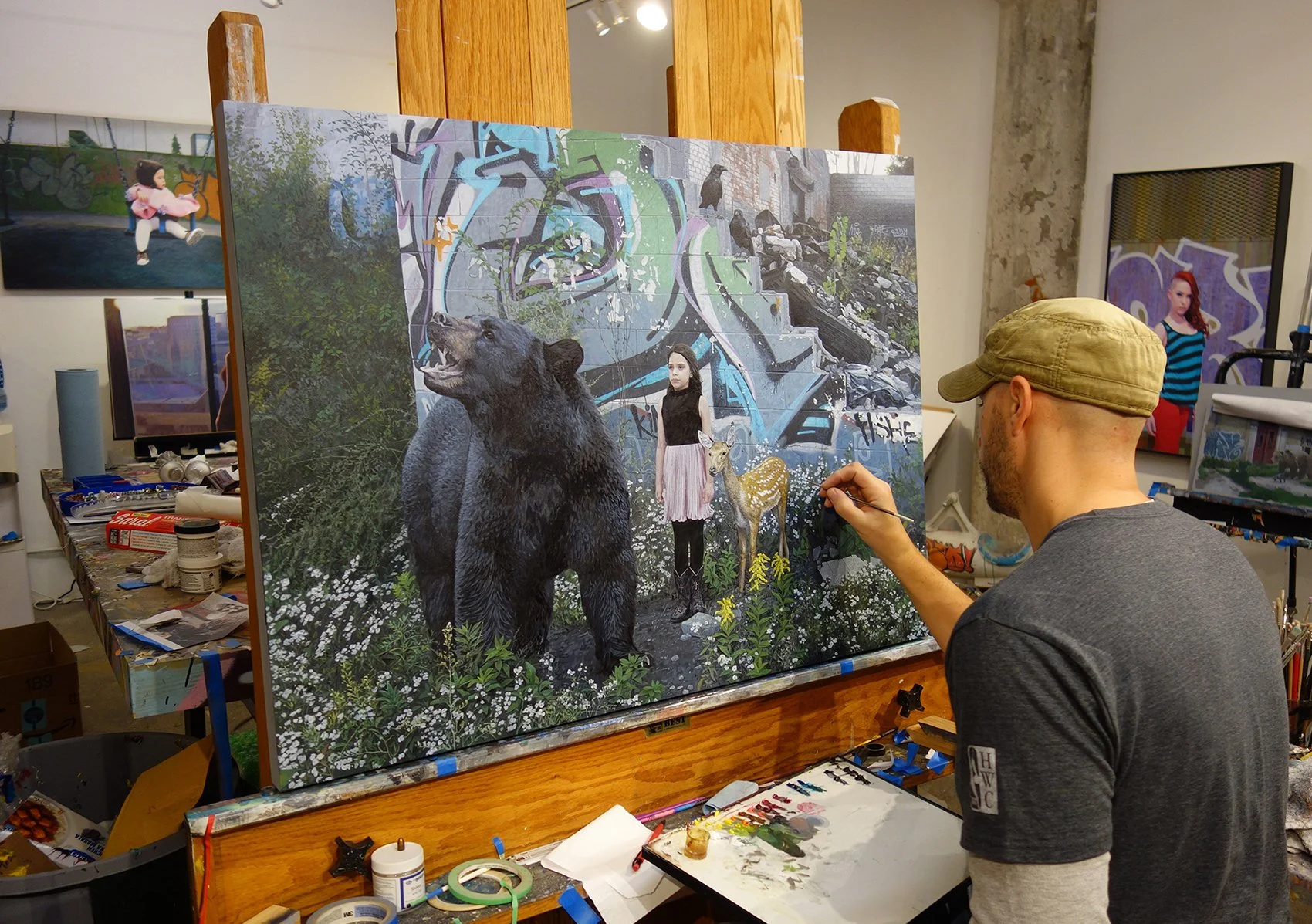 An artist painting a large, detailed mixed-media piece on a canvas, featuring a girl with dark hair, a large bear, a deer, and graffiti-style background elements, inside an art studio with other artworks visible.