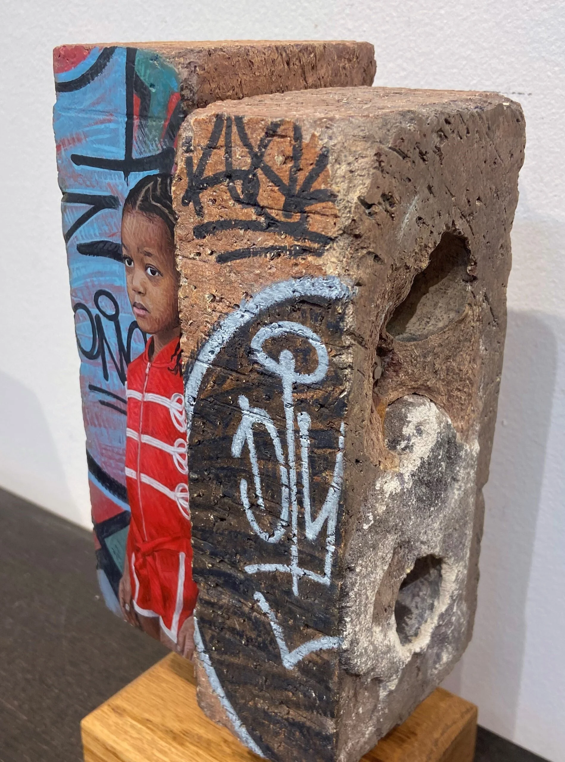 A mixed-media art piece featuring a portrait of a young girl with braided hair and a red jacket, painted on a brick with graffiti-style markings and a white painted peace symbol.