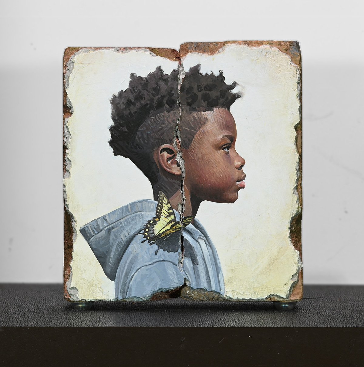 A painting on a cracked, rectangular canvas of a young boy in profile with an afro hairstyle, a yellow butterfly on his shoulder, and a hoodie.
