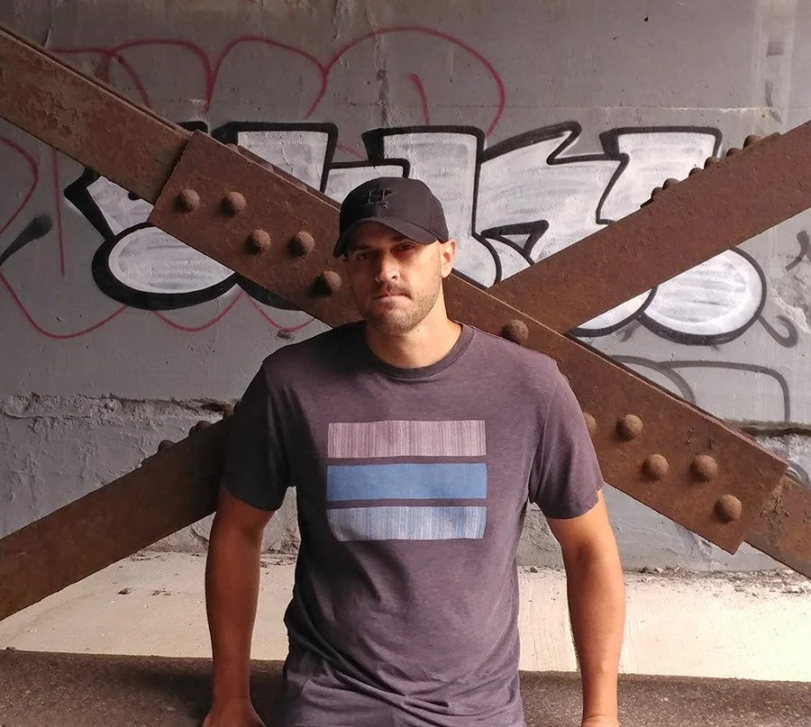 A man wearing a black cap and dark T-shirt standing in front of a graffiti-covered wall with a rusted metal beam crossing diagonally behind him.