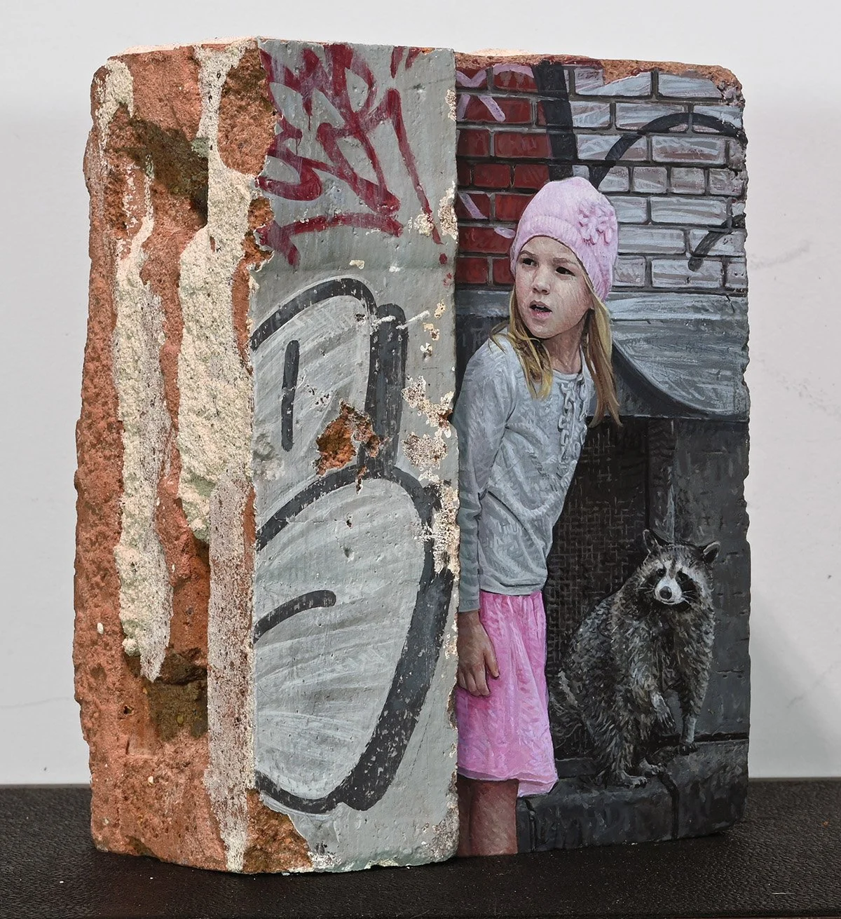 A mixed media artwork that combines a piece of brick wall with a painted mural of a young girl wearing a pink beanie, a gray sweater, and pink skirt, standing next to a raccoon painted on the wall.