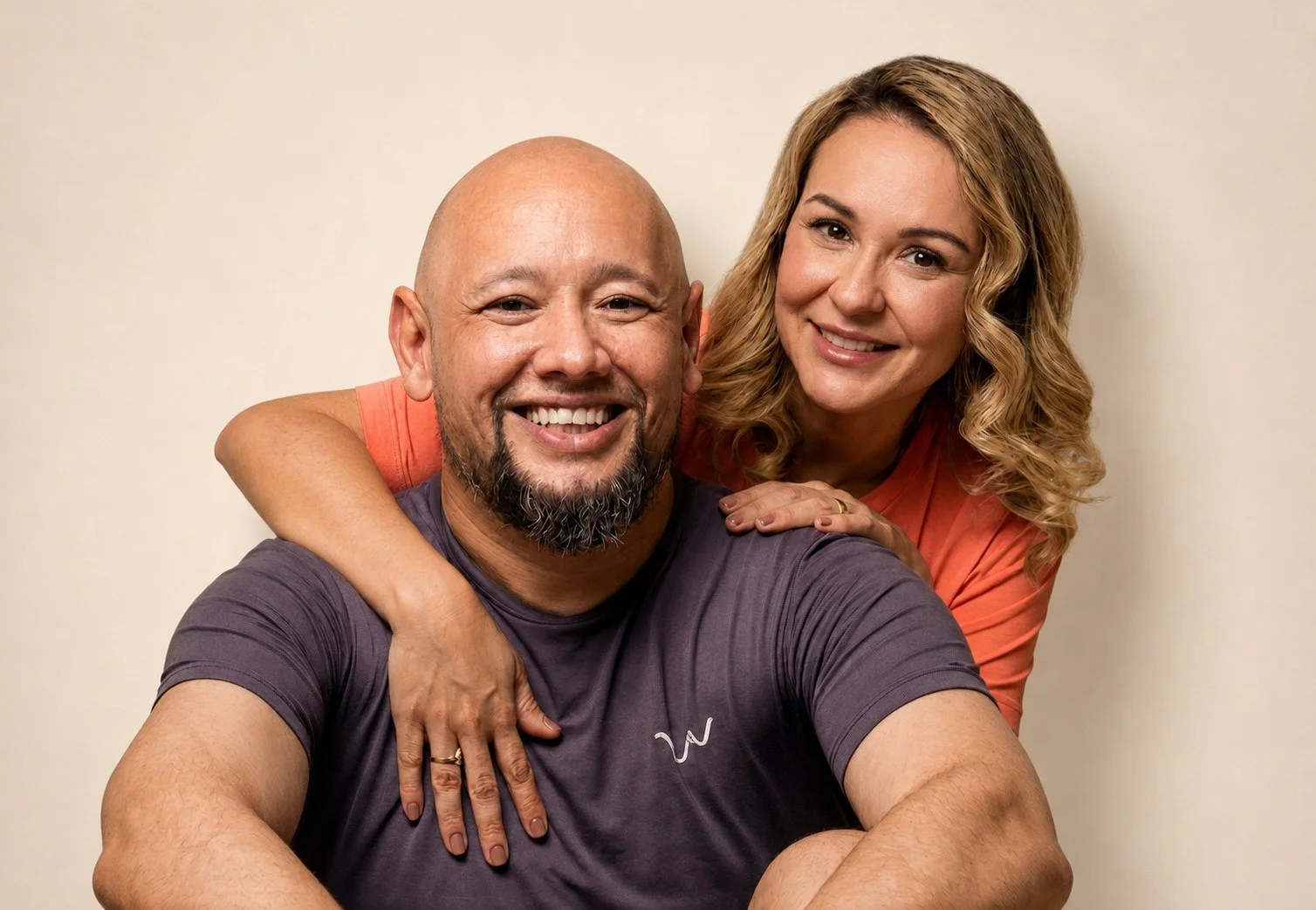 Homem e mulher sorrindo, mulher com braço no ombro do homem, fundo bege.