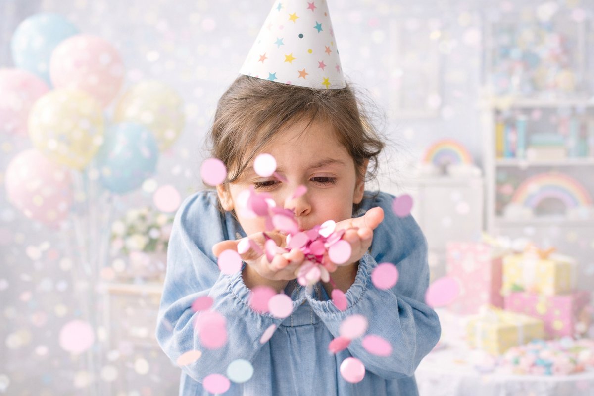 Ein Kind trägt eine Geburtstagsmütze und bläst Konfetti in die Luft bei einer Geburtstagsfeier im Raum, der mit bunten Ballons und Geschenken dekoriert ist.