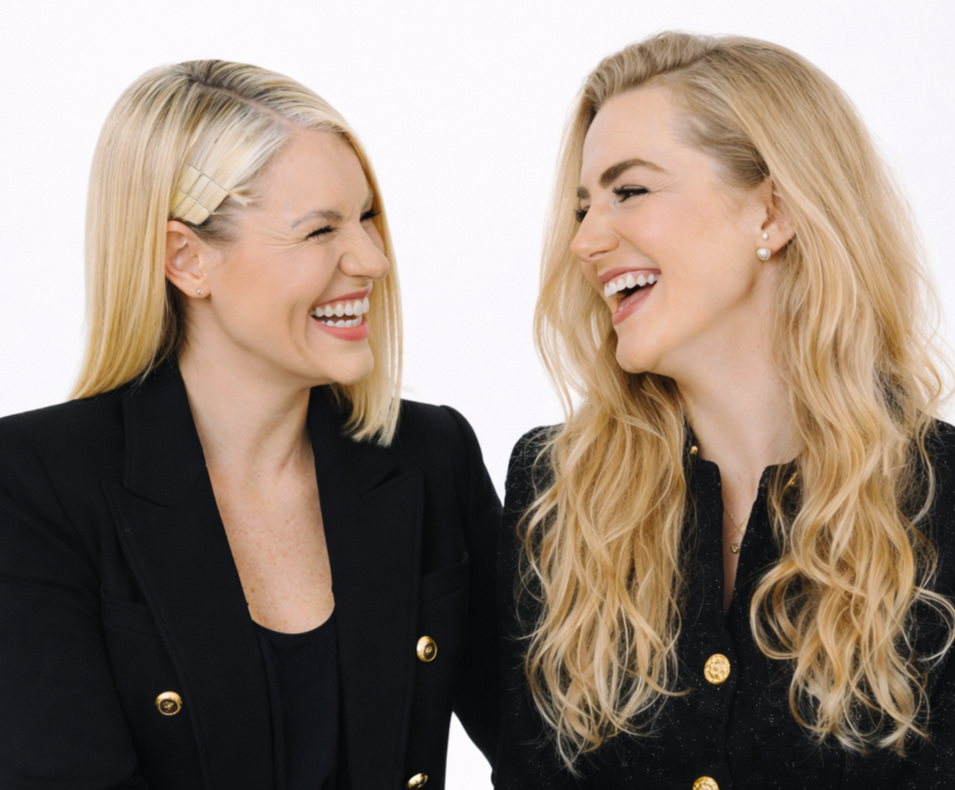 Zwei lachende Frauen in schwarzen Blazern, die miteinander lachen, vor einem weißen Hintergrund.