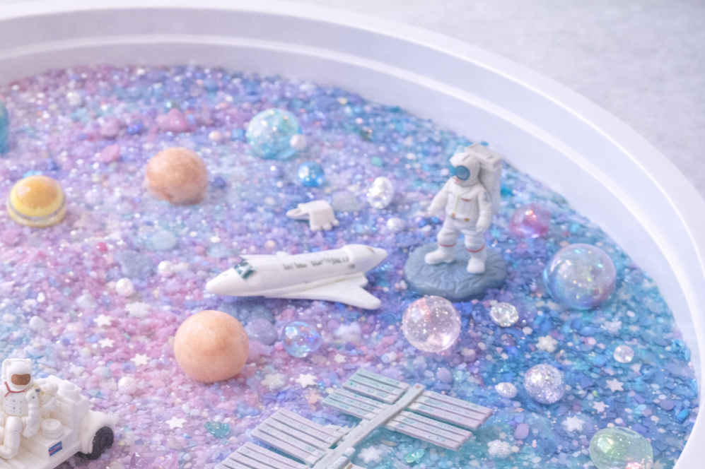 Miniaturmodell einer Weltraumlandschaft mit Astronauten, Raumfahrzeugen, bunten Glitzer- und Perlenkugeln, Sternchen und Weltraumobjekten auf einem randabschlossenen weißen Behälter.