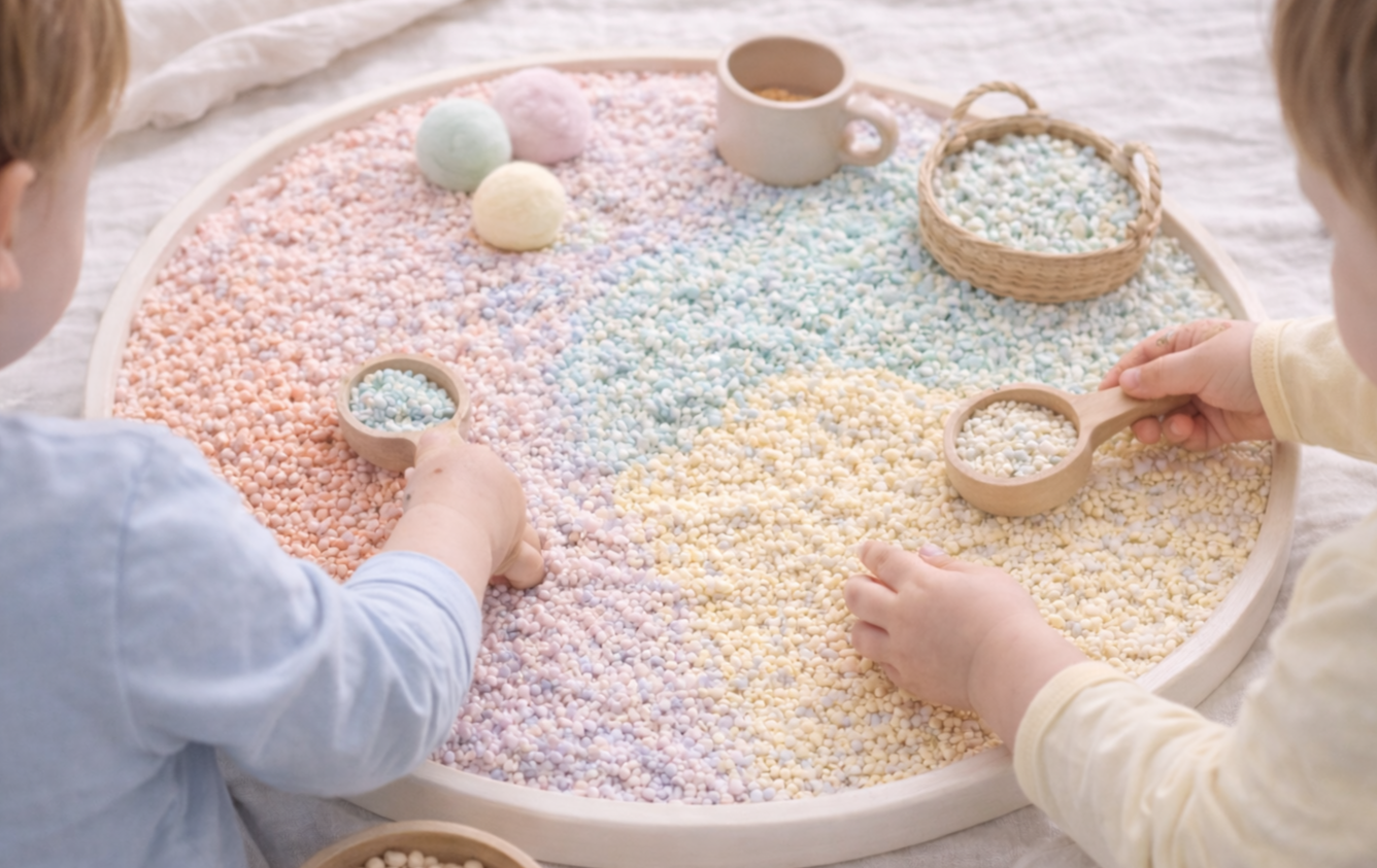 Zwei Kinder spielen mit bunten Maiskörnern in einem großen runden Spielteller, der auch ein Tasse und eine kleine Schüssel enthält.