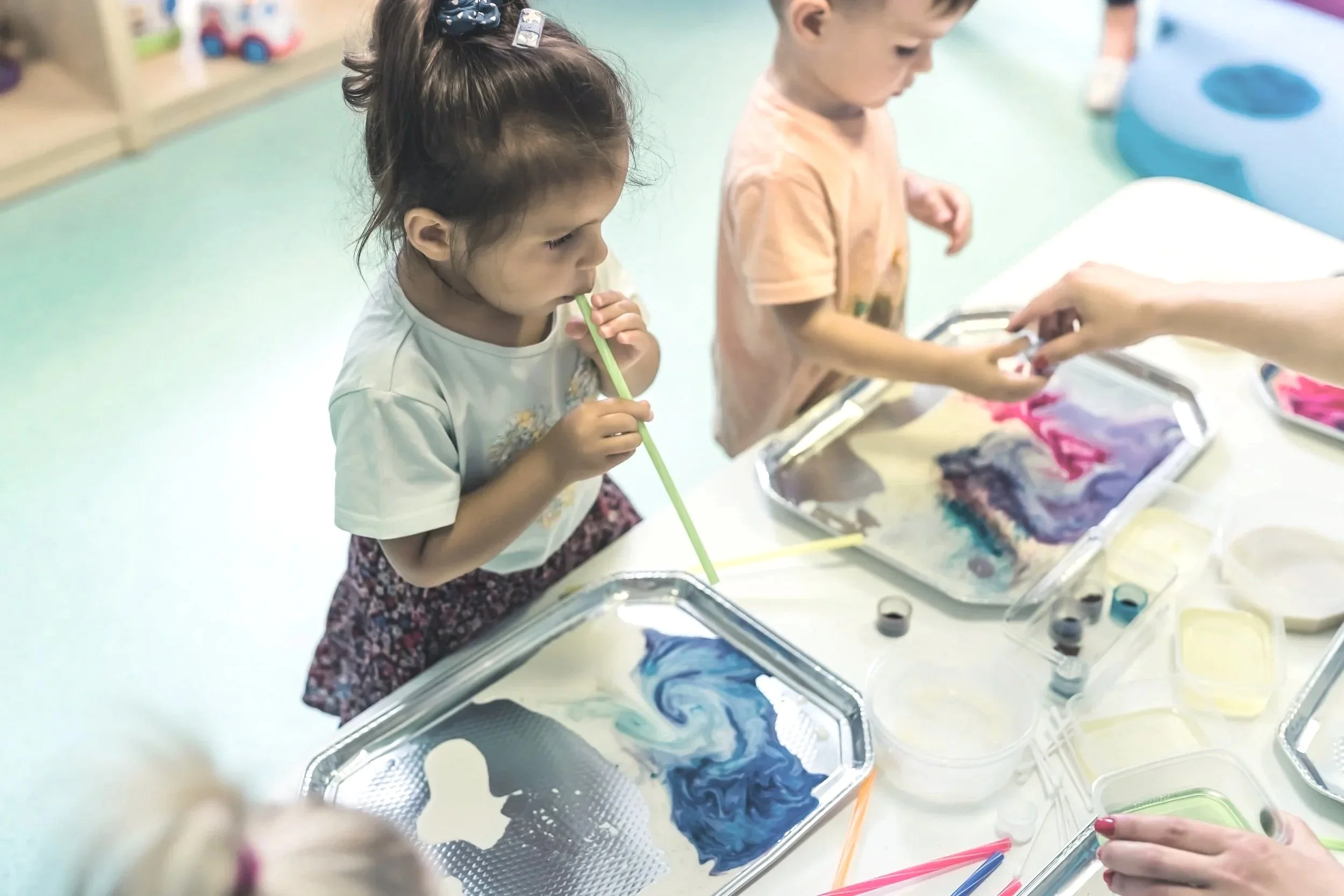Zwei Kinder, ein Mädchen und ein Junge, experimentieren mit bunten Farben und Rührstäbchen auf Metalltabletts in einem Kunst- oder Bastelraum.