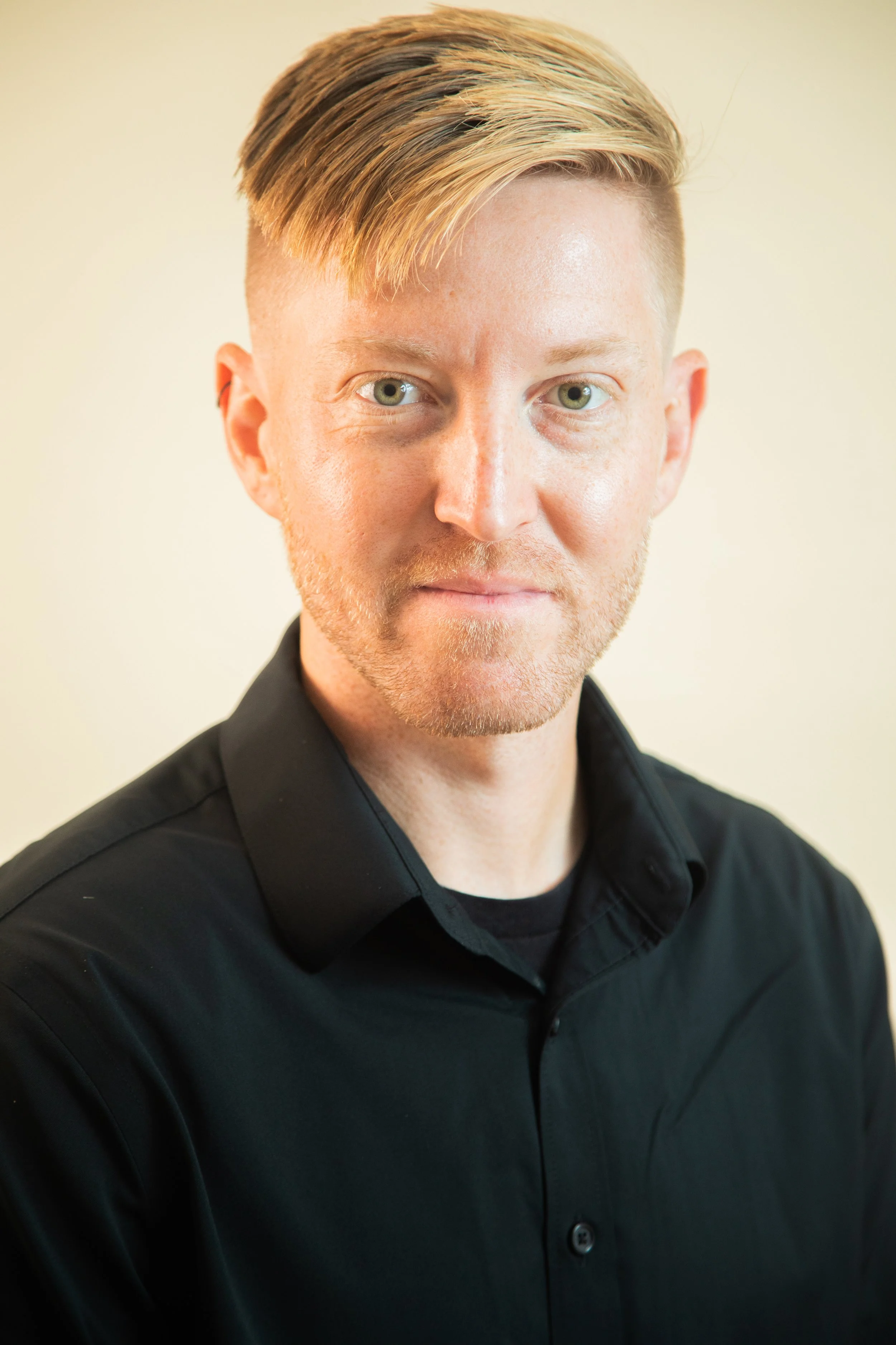 A young man with blond hair styled to the side, wearing a black collared shirt, looking directly at the camera with a neutral expression, against a light, neutral background.
